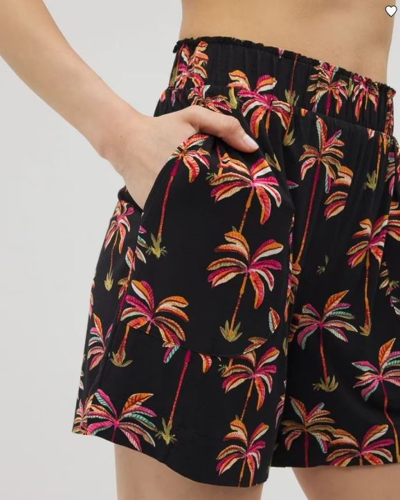 Close-up of a person wearing black shorts with colorful palm tree print, hand in pocket, white background.