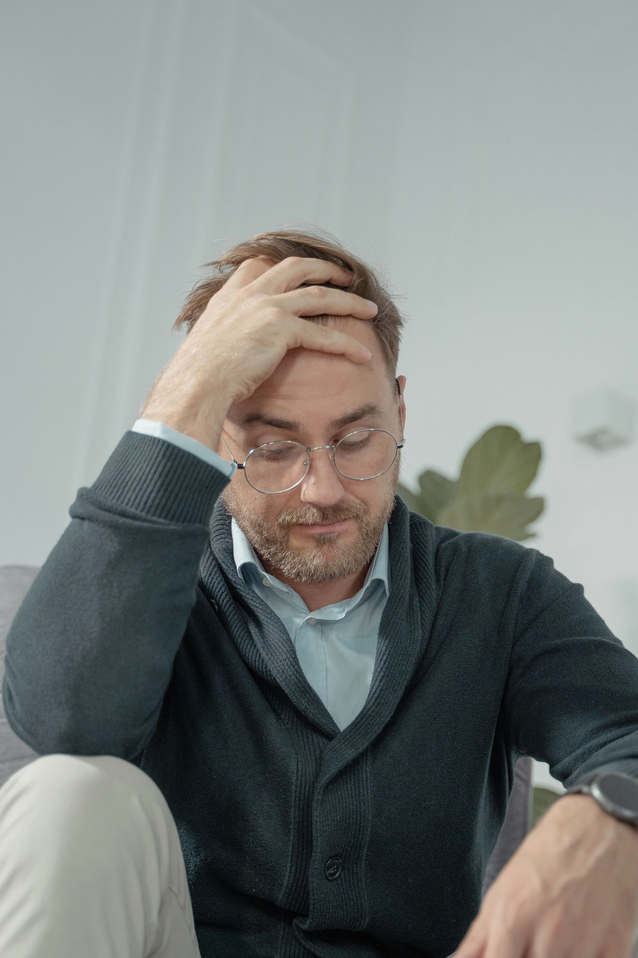 A thoughtful man with glasses sits indoors, hand on head, reflecting stress or contemplation