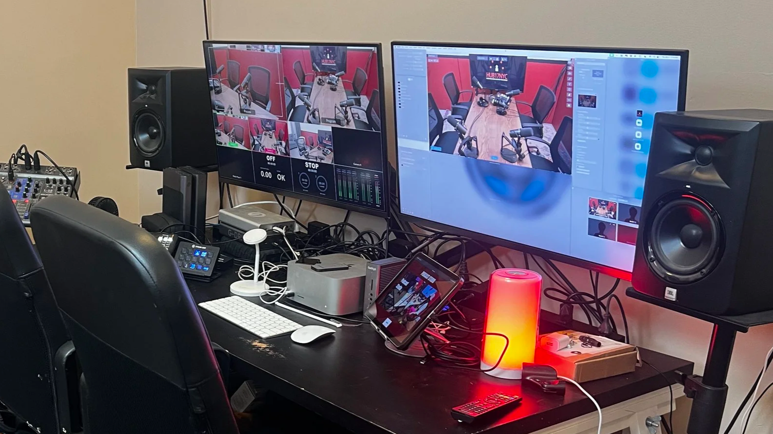 A recording or streaming setup with dual computer monitors displaying a video conference or recording session, surrounded by audio speakers, a mixing console, a tablet, a keyboard, a mouse, and various cables on a black desk. There's a red cylindrical lamp, a small white lamp, and other electronic devices in the workspace.