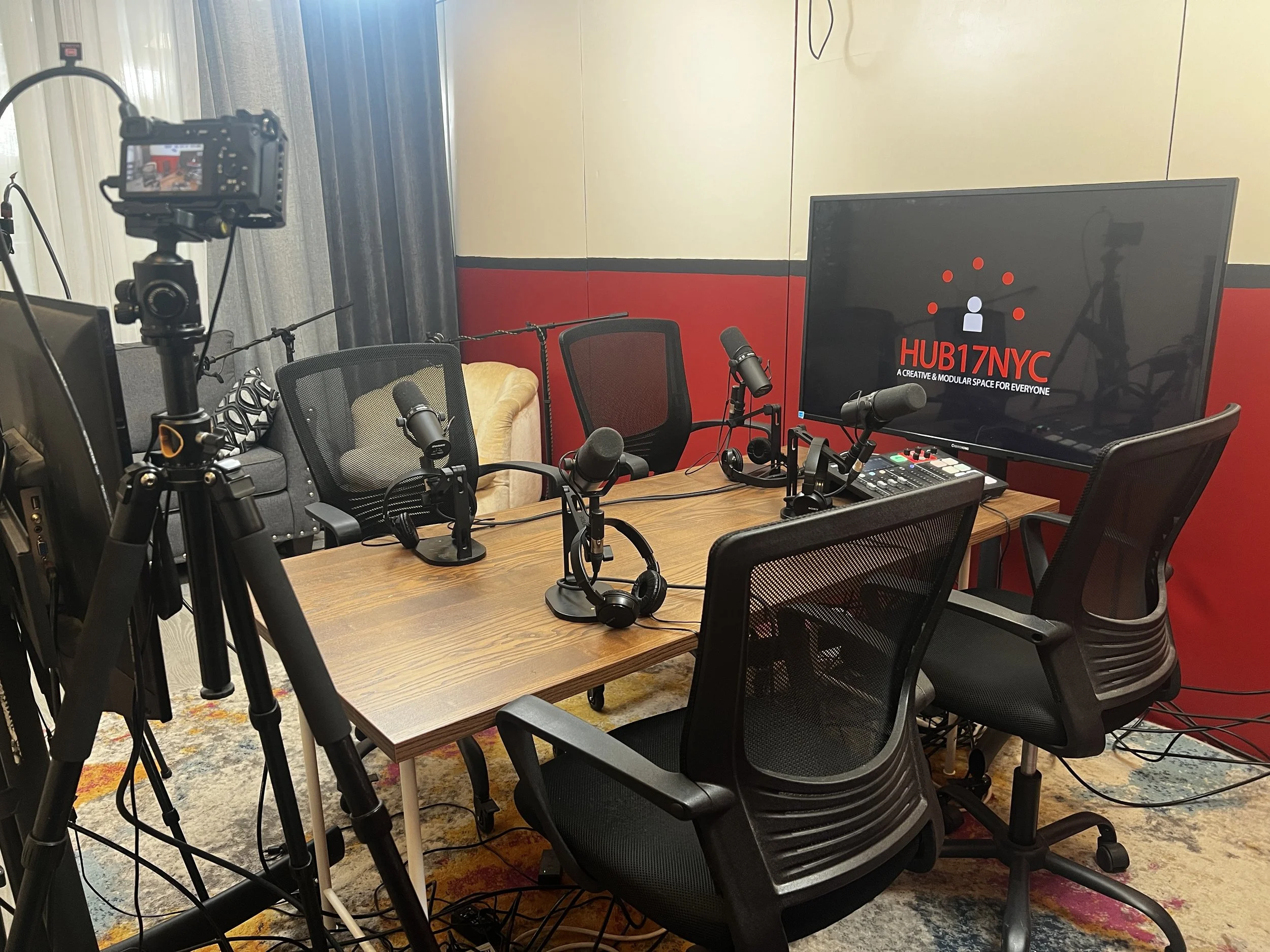 A recording studio setup with a wooden table, microphones, headphones, and a large monitor displaying 'HUB17NYC' logo and text about a creative space, surrounded by black office chairs and filmed equipment.