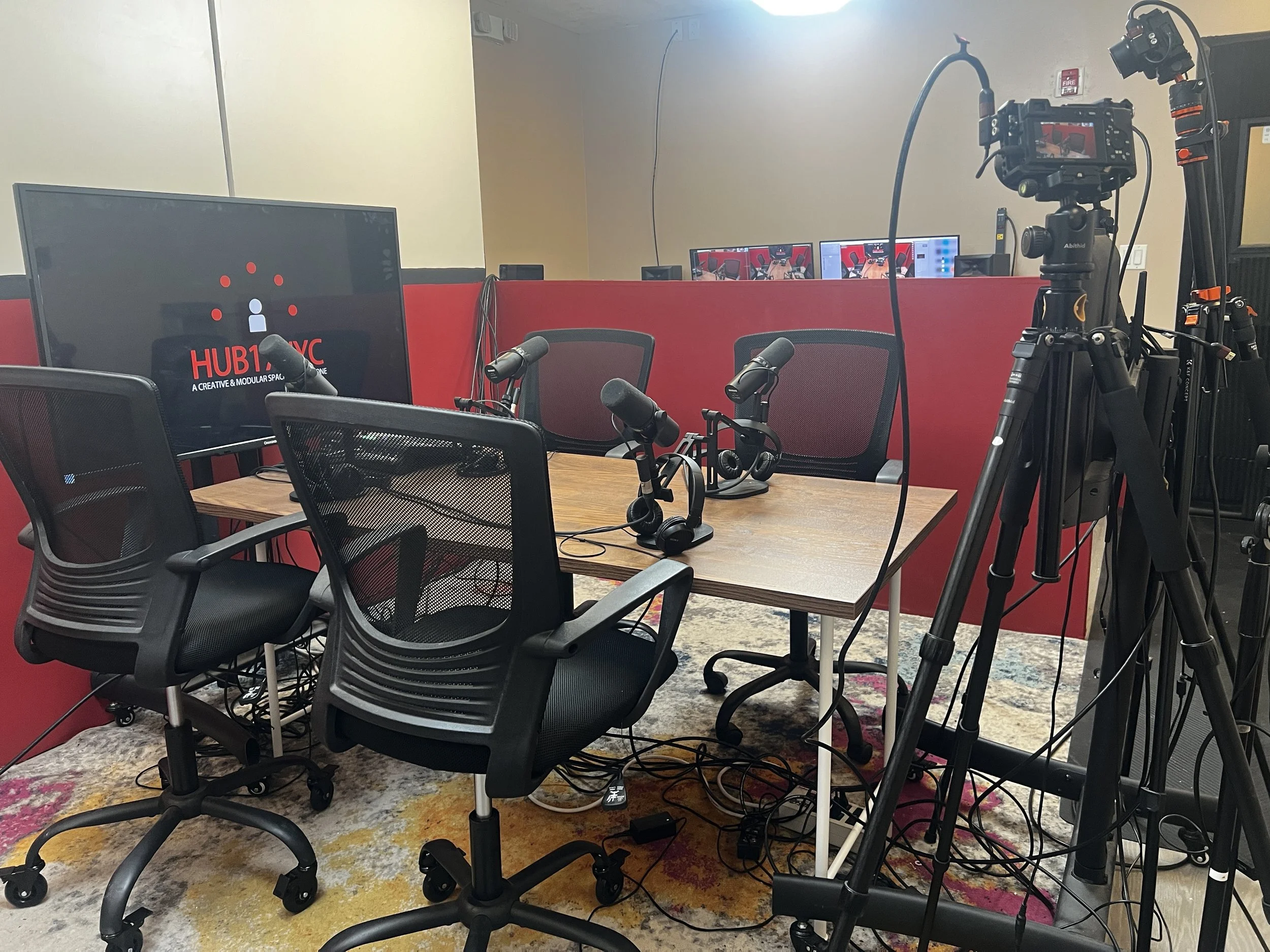 A recording studio or podcast room setup with multiple microphones on a table, a camera on a tripod, a large monitor displaying 'HUB17NYC' and some chairs, with dual computer monitors in the background, and a colorful rug on the floor.