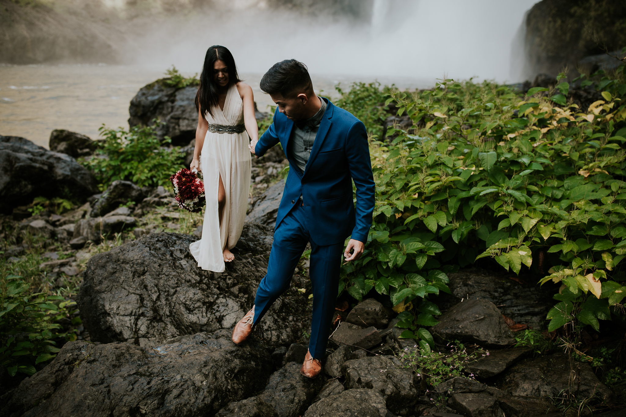 Groom leading adventurous bride over rocks in front of Snoqualmie falls