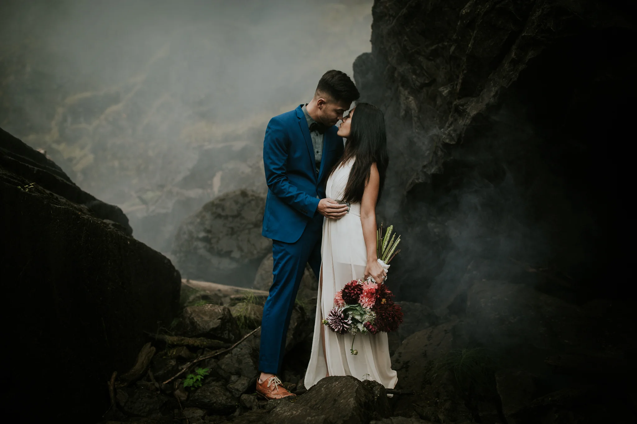 Bride and groom embracing in front of black rocks with mist near waterfall