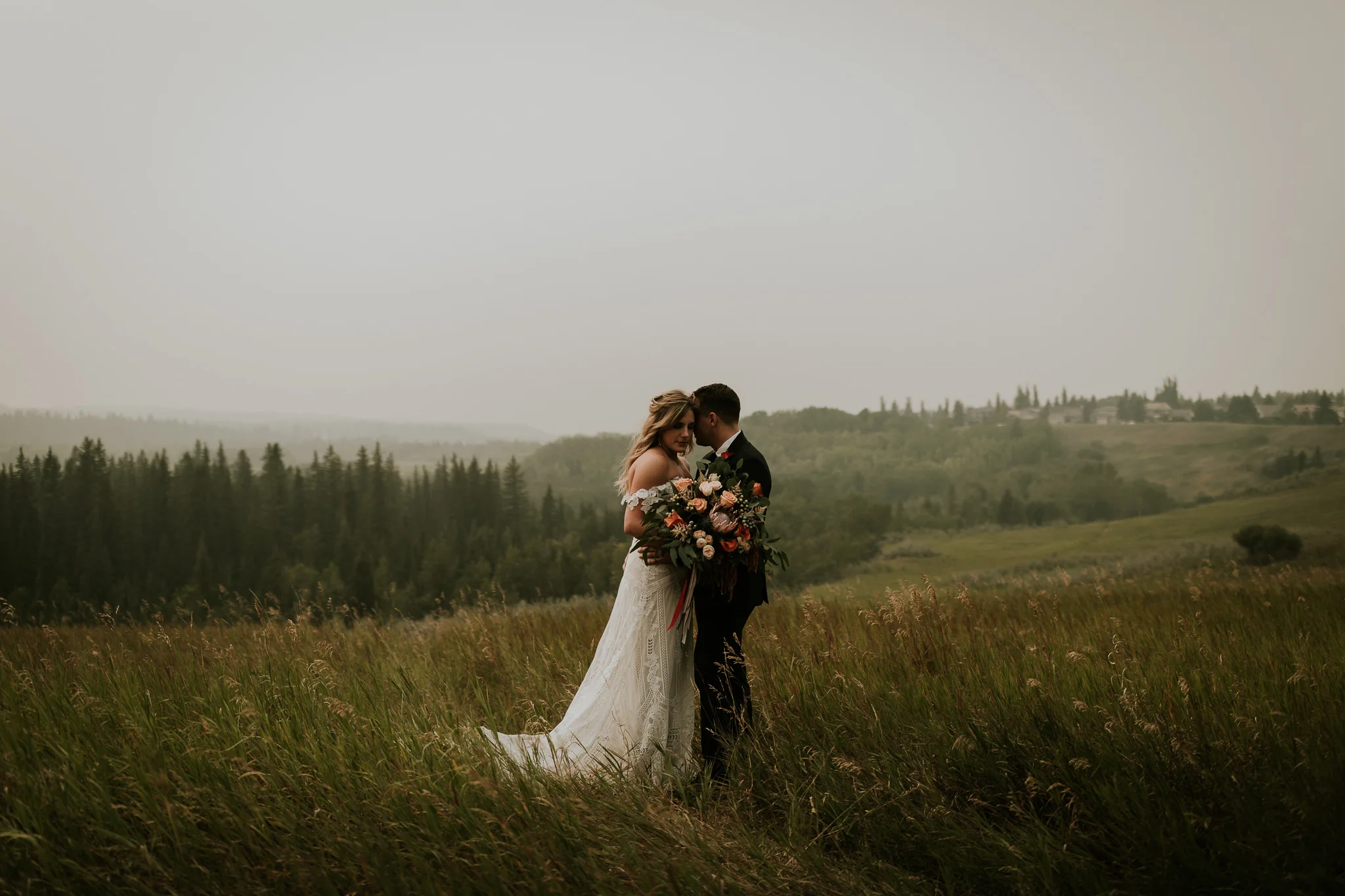 Romantic wedding photo of boho couple wearing Rue de Seine bridal gown and holding floral bouquet, embracing her husband in Fish Creek Park Calgary