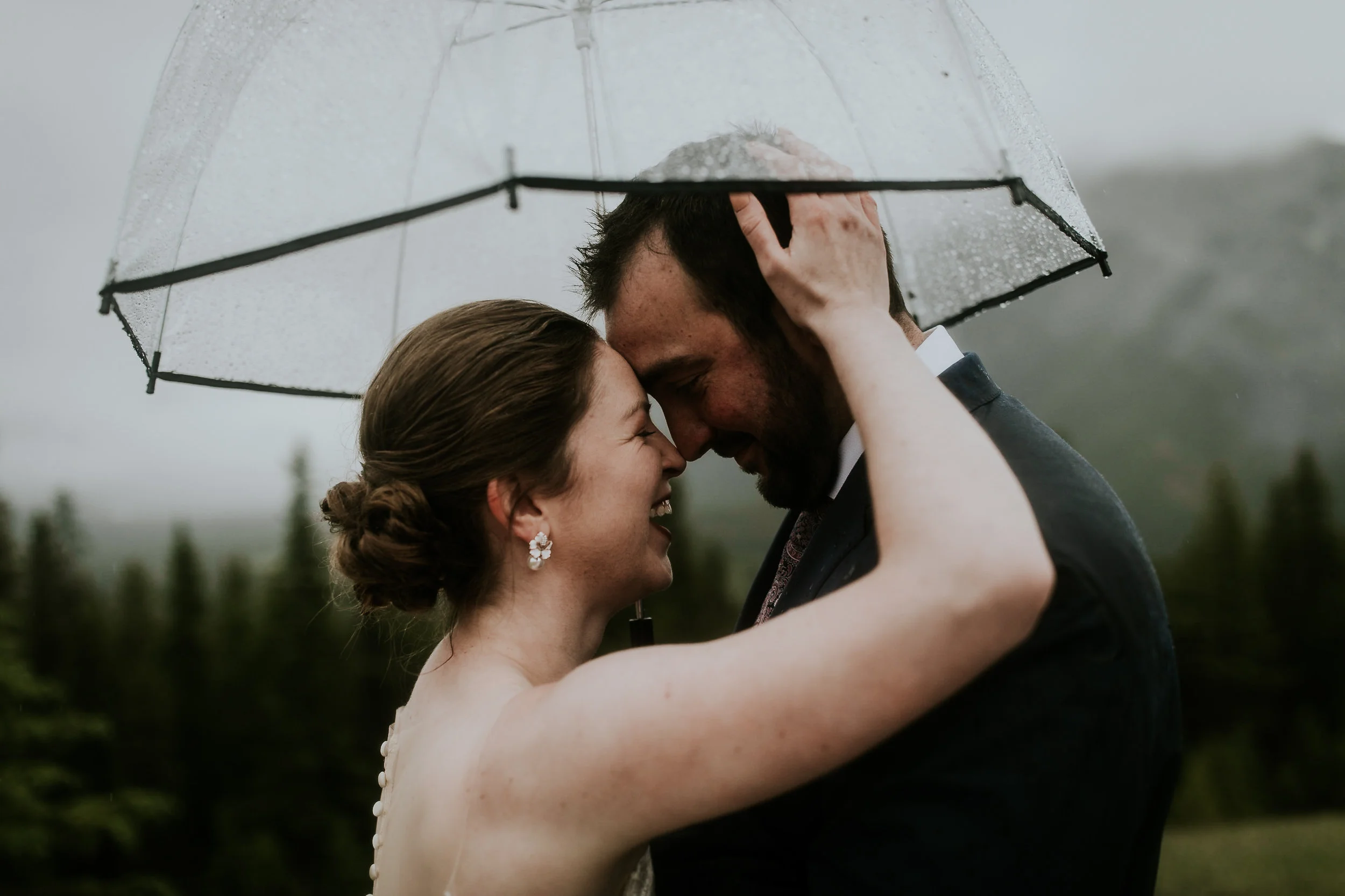  bride and groom holding each others heads and smiling and crying as rain pours under umbrella during destination wedding  