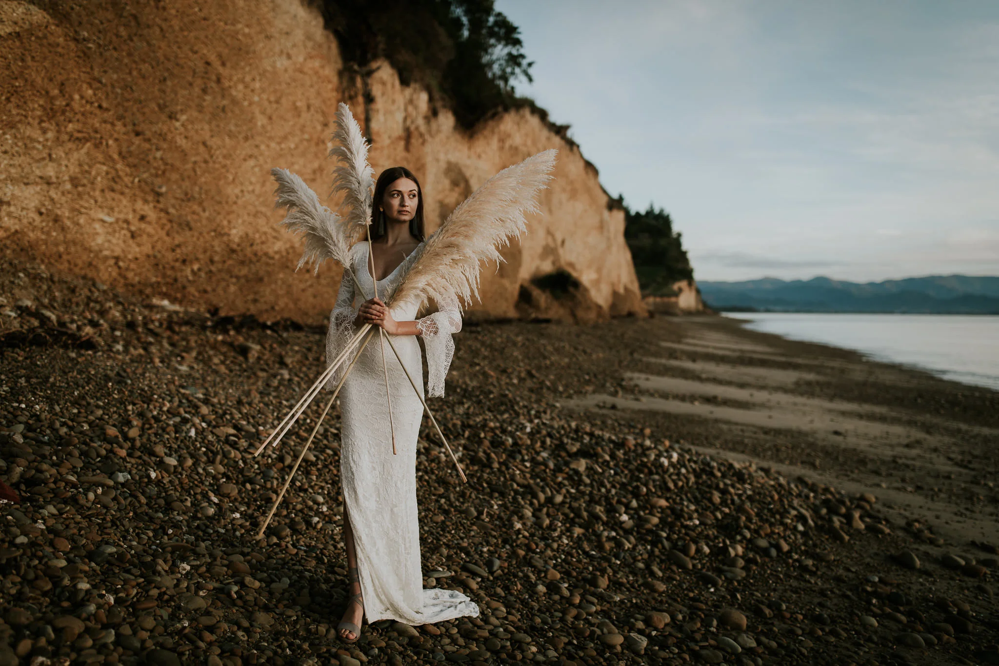  new zealand bride holds pampas grass and looks out to ocean as giant cliffs light up in the background at sunrise during destination wedding  