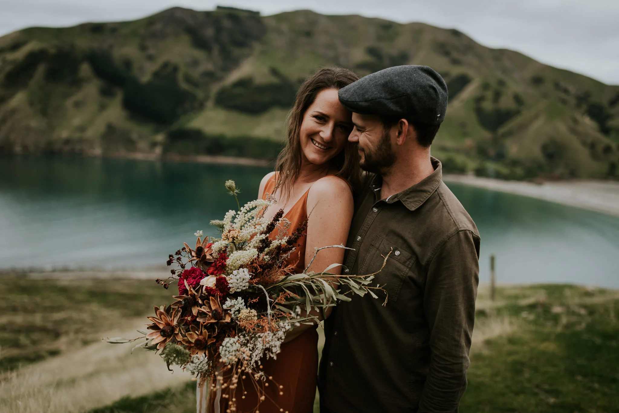  couple holding large bouquet of flowers smiling and laughing looking at each other with new zealand beach in the background during destination elopement 