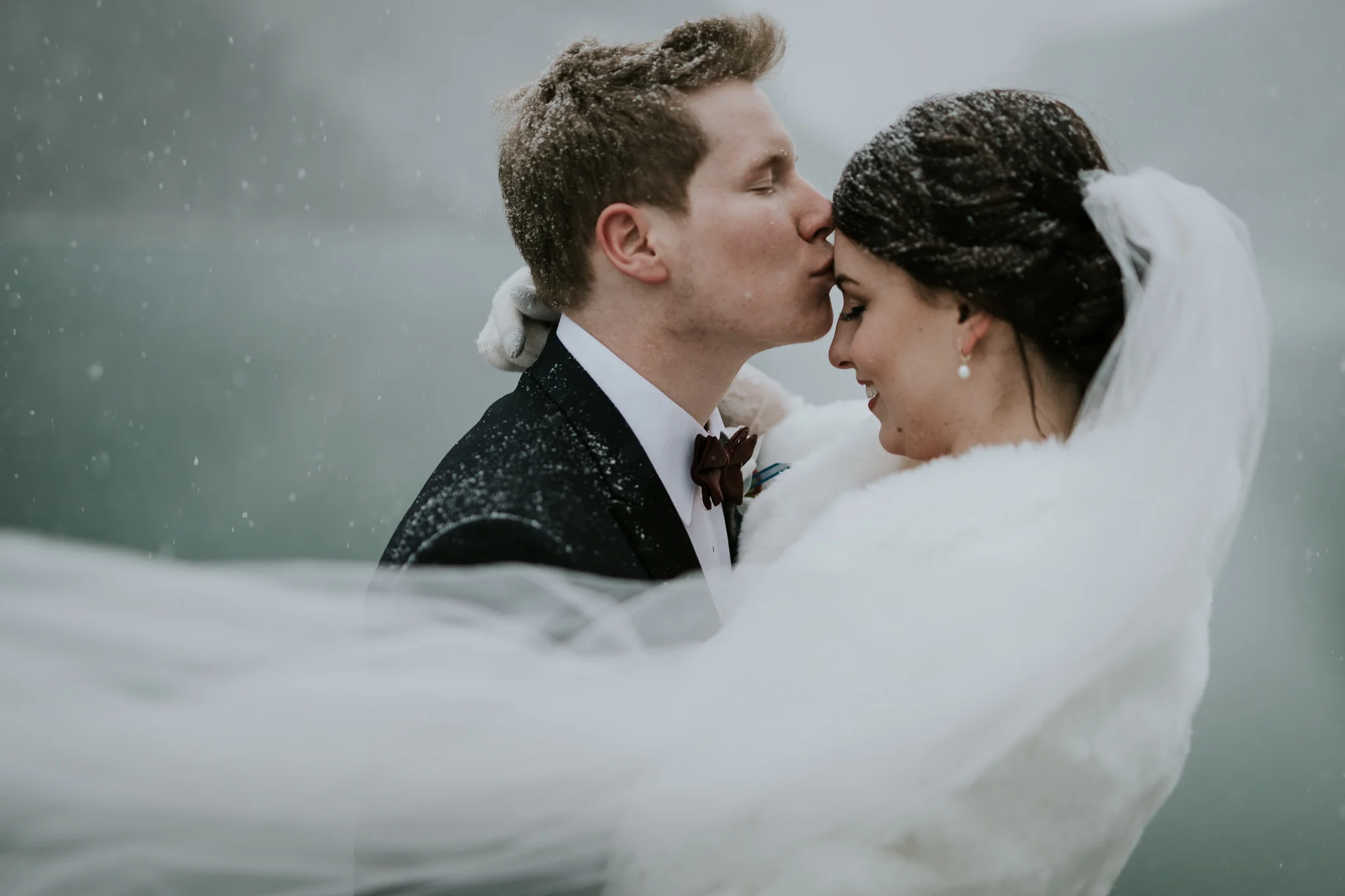  groom kisses brides forehead as veil blows across during snowy winter portraits during destination wedding in Canada 