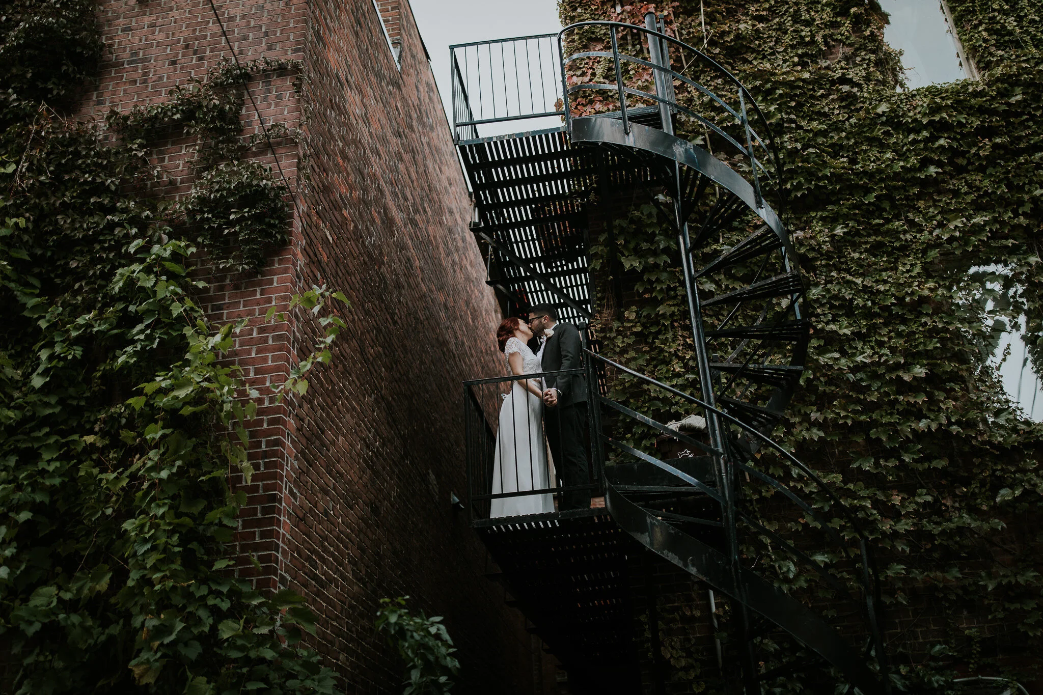  newly married couple embrace on spiraling staircase with brick walls covered in vines in montreal during destination wedding  