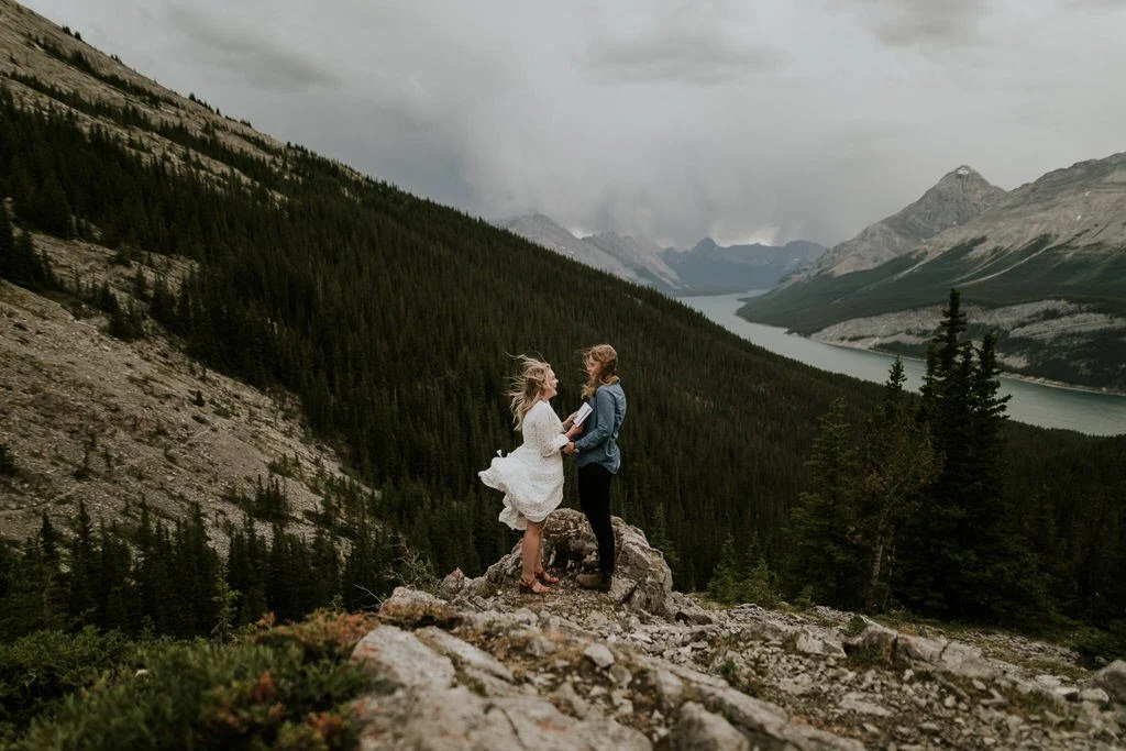  two brides eloping on top of mountain with view of mountains and a lake in the background as wind blows during destination elopement 