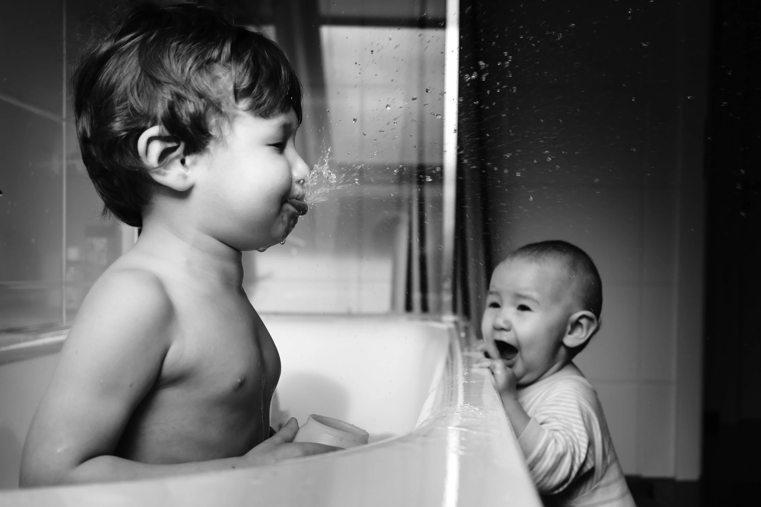 Black and white photo with boy spitting water in the foreground and baby in background laughing