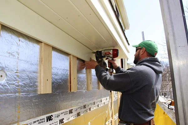 Fenton nails the frieze board to the wall strapping. The frieze board runs around the entire house just below the plane of the roof eaves and the dormer windows, serving as a unifying trim element that ties the home together visually.