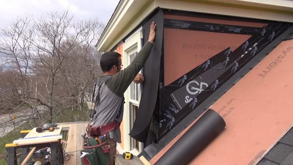 Pollard applies asphalt felt paper over the ZIP sheathing, sliding the top edge of the paper up underneath the unstapled lower edge of a strip of paper already applied to the eave under the trim board. To his left, Pollard tucks the paper under Mortairvent rainscreen fabric already installed under the window trim.