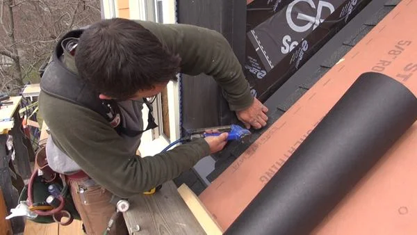 Pollard fastens the bottom edge of the asphalt felt paper over the previously applied self-adhering bituminous membrane flashing that covers the top edge of the aluminum step flashing.