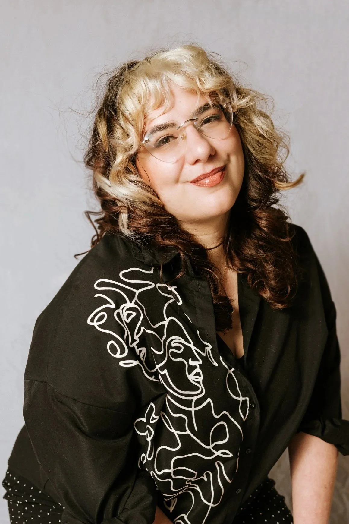 A woman with curly blonde and brown hair, wearing glasses, a black shirt with a white line art face design, and black pants with white polka dots, smiling at the camera.