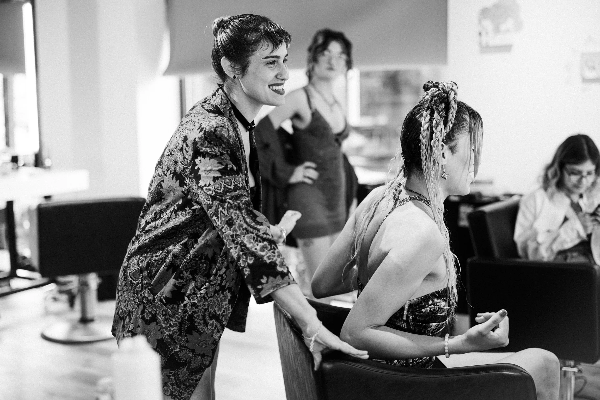 A woman with short dark hair and tattoos on her arms smiling while talking to a seated woman with braided hair, wearing a tank top, in a room with other people in the background.