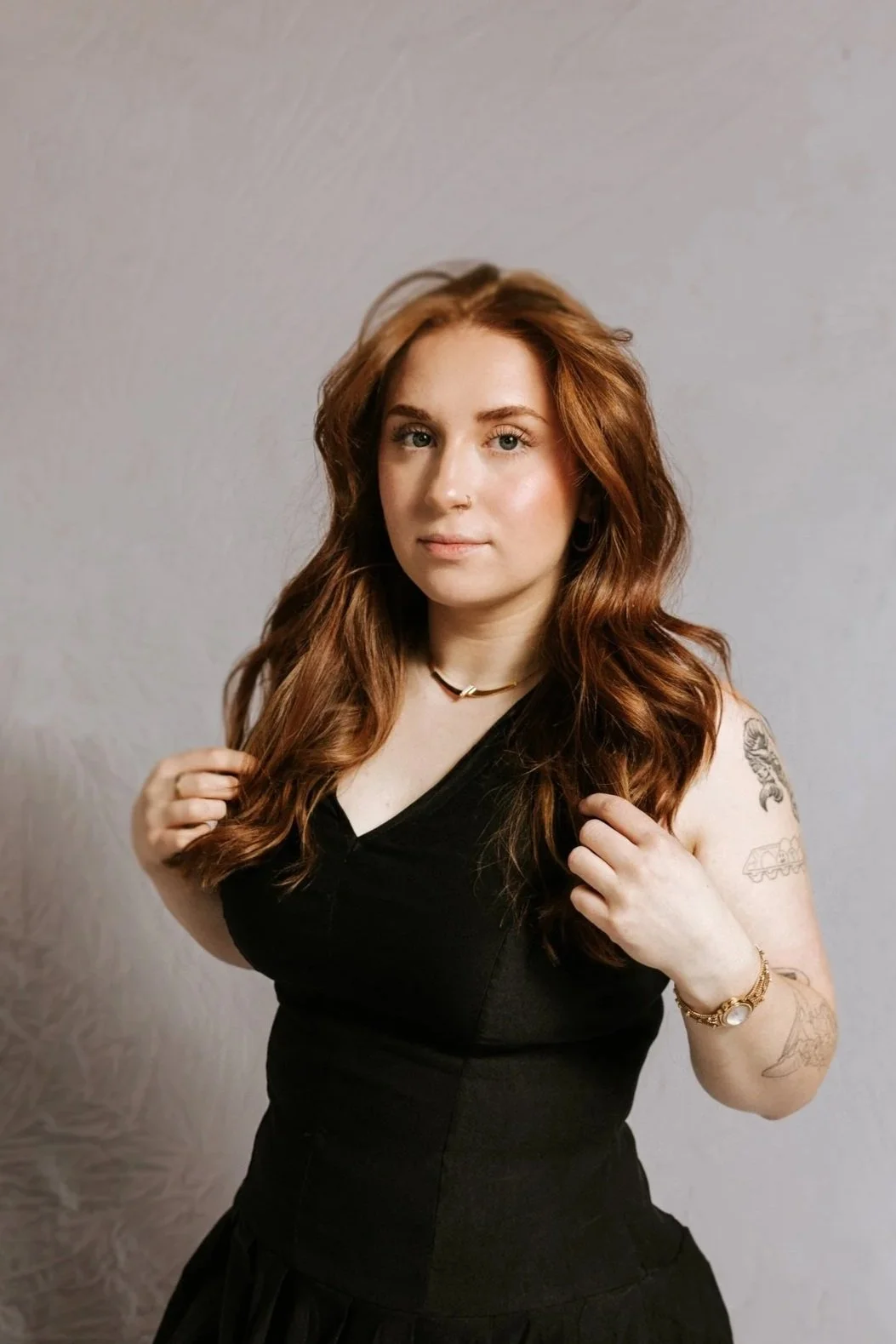 A woman with long red hair, wearing a black sleeveless top, jewelry, and tattoos on her right arm, standing against a plain light-colored background.