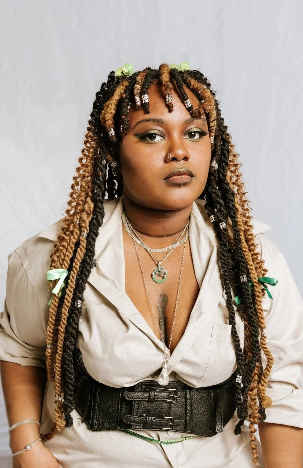 A woman with dreadlocks and multiple necklaces, wearing a beige shirt and black wide belt.