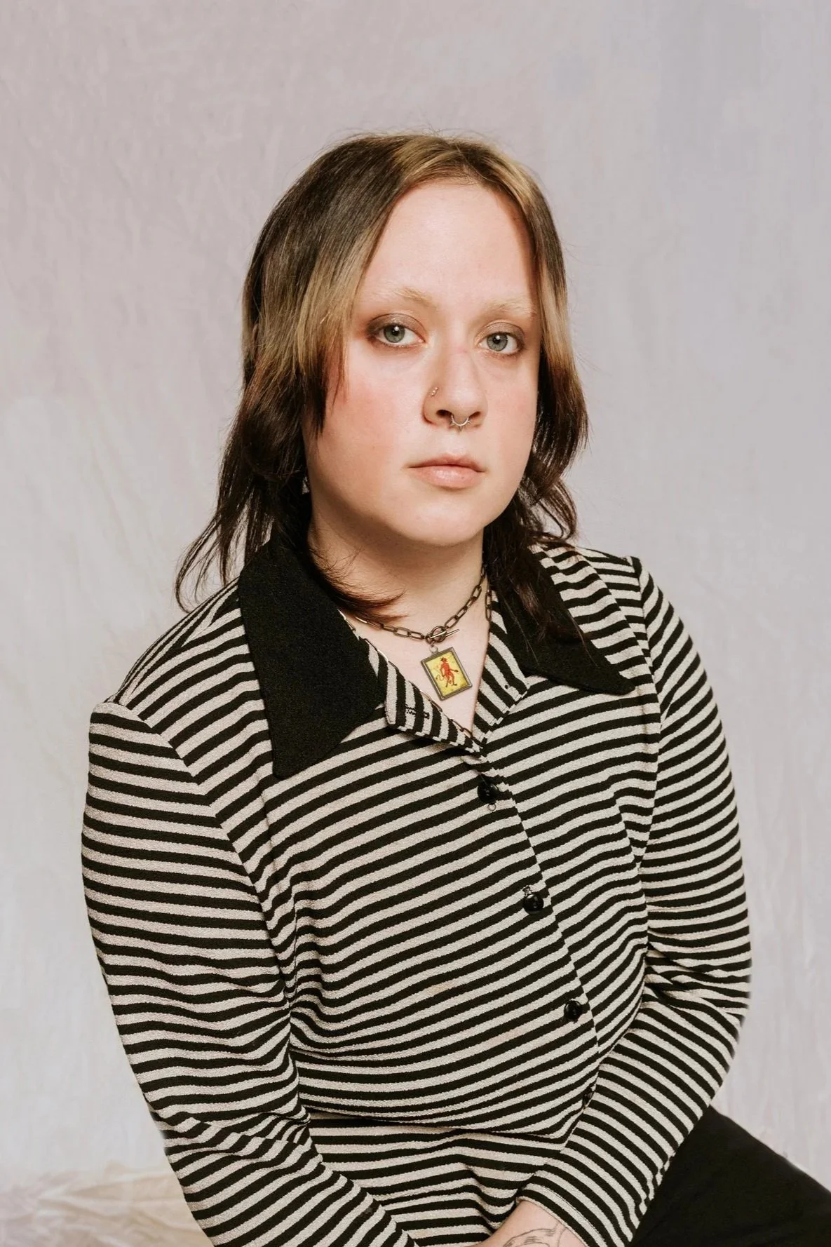 A young woman with shoulder-length brown hair and light skin, wearing a black and white striped shirt with black collar and buttons, a necklace with a yellow pendant, and a nose ring, sitting against a neutral background.