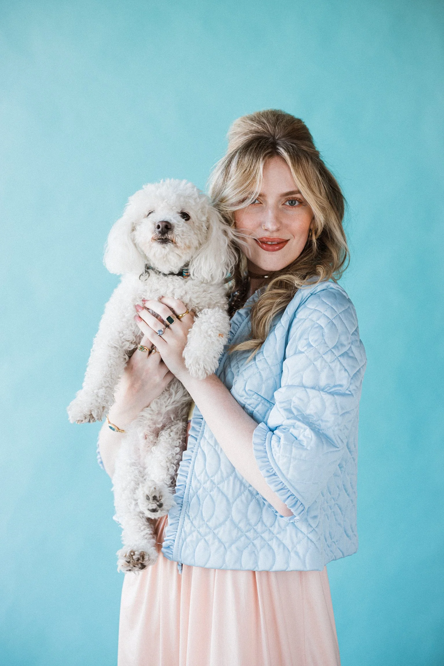 A woman with blonde hair wearing a pink dress and blue quilted jacket holding a white poodle dog against a light blue background.