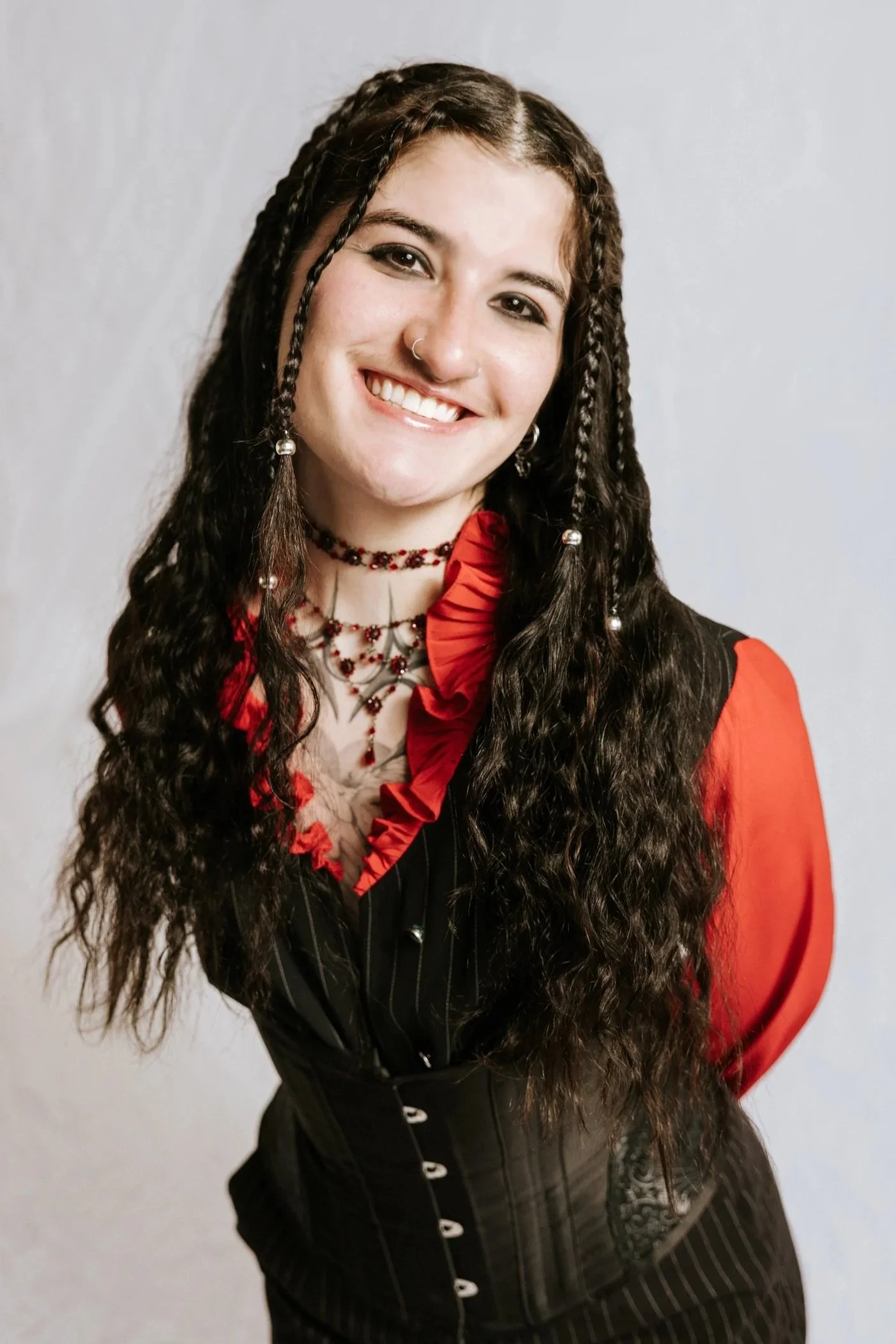 A young woman with long, curly hair styled with braids, wearing a nose ring, multiple necklaces, a red and black blazer, and smiling against a plain background.