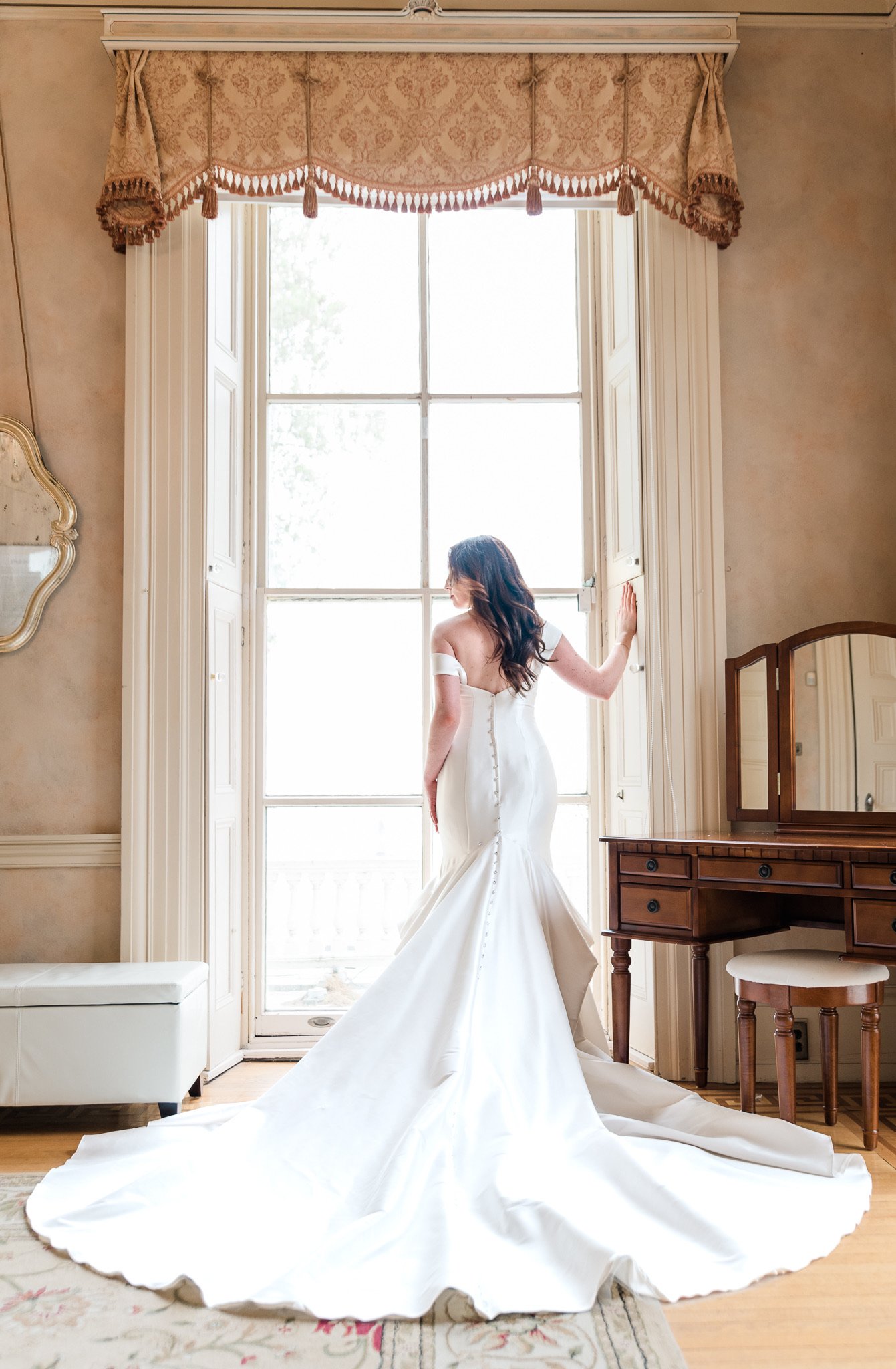 A woman in a white wedding dress stands by a large window with her back to the camera, in a room with vintage furniture and curtains.