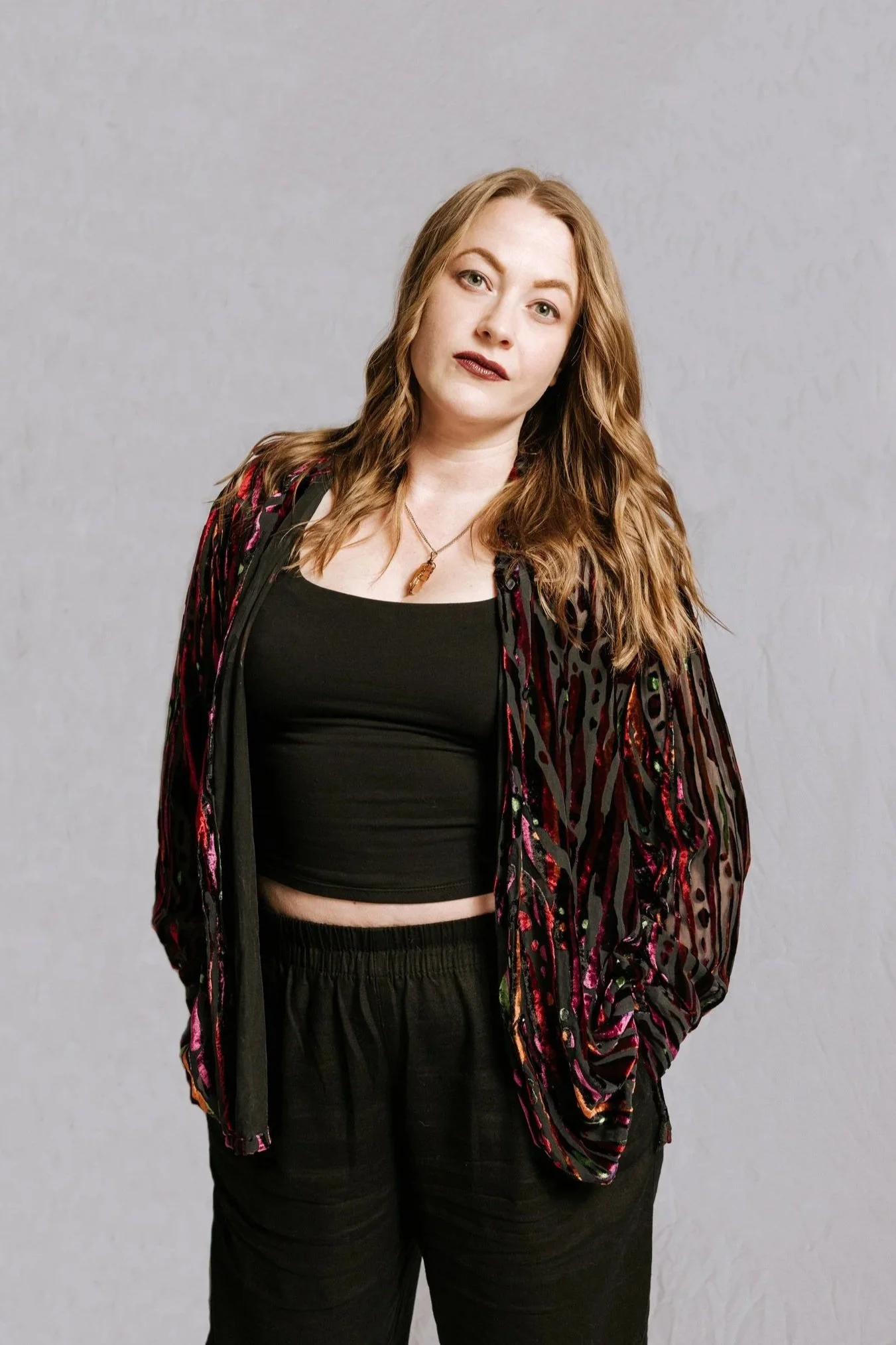 Red-haired woman wearing a black crop top, black pants, and a colorful, patterned jacket, standing against a plain gray background.