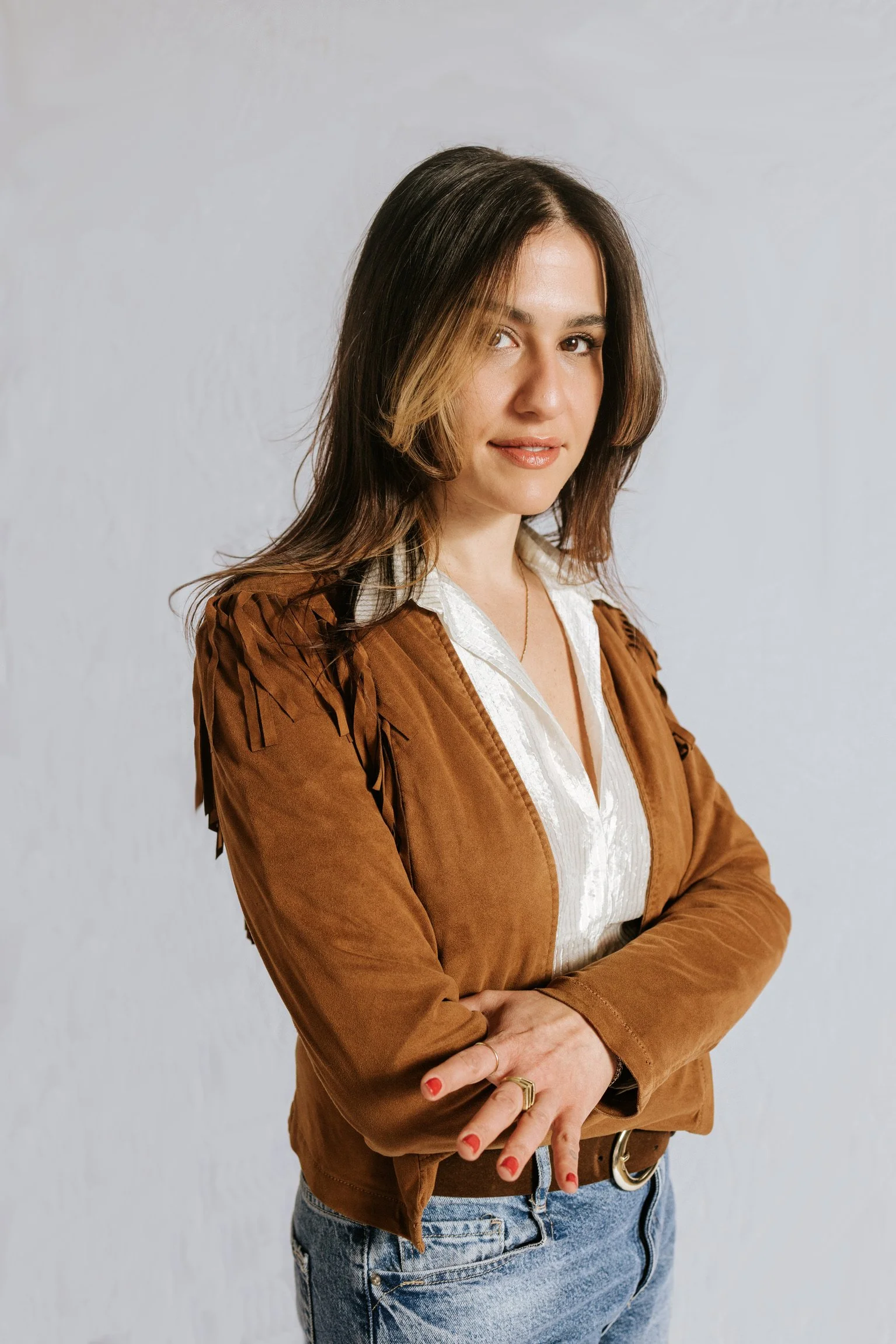 A person with shoulder-length brown hair and light skin, wearing a white blouse, a brown jacket, and light blue jeans, stands with arms crossed against a plain light grey background.