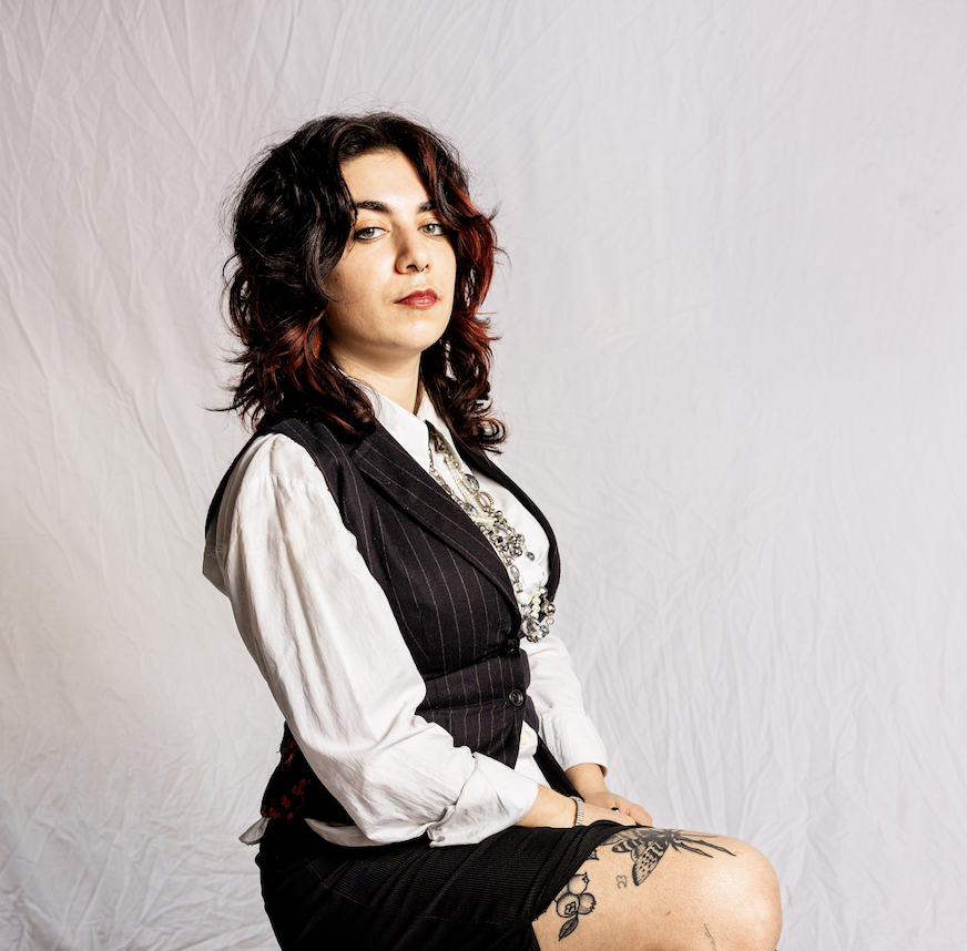 A woman with dark wavy hair and red highlights sitting on a chair against a plain backdrop, wearing a white blouse, a black pinstripe vest, and a skirt, with tattoos on her thigh, and accessorized with necklaces.
