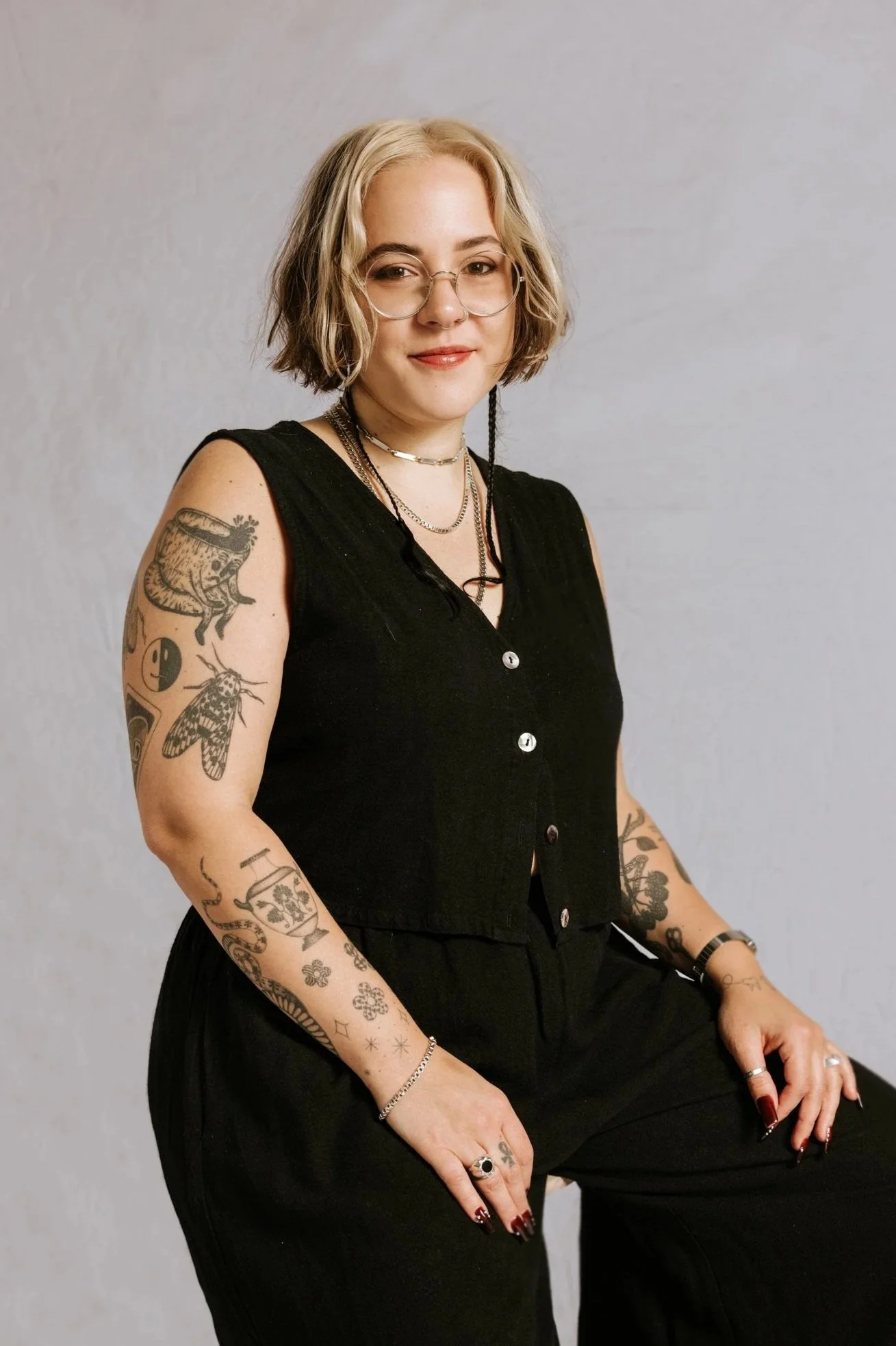 A person with short blonde hair, glasses, and multiple tattoos on her arms, wearing a black sleeveless top and jewelry, sitting against a plain light-colored wall.