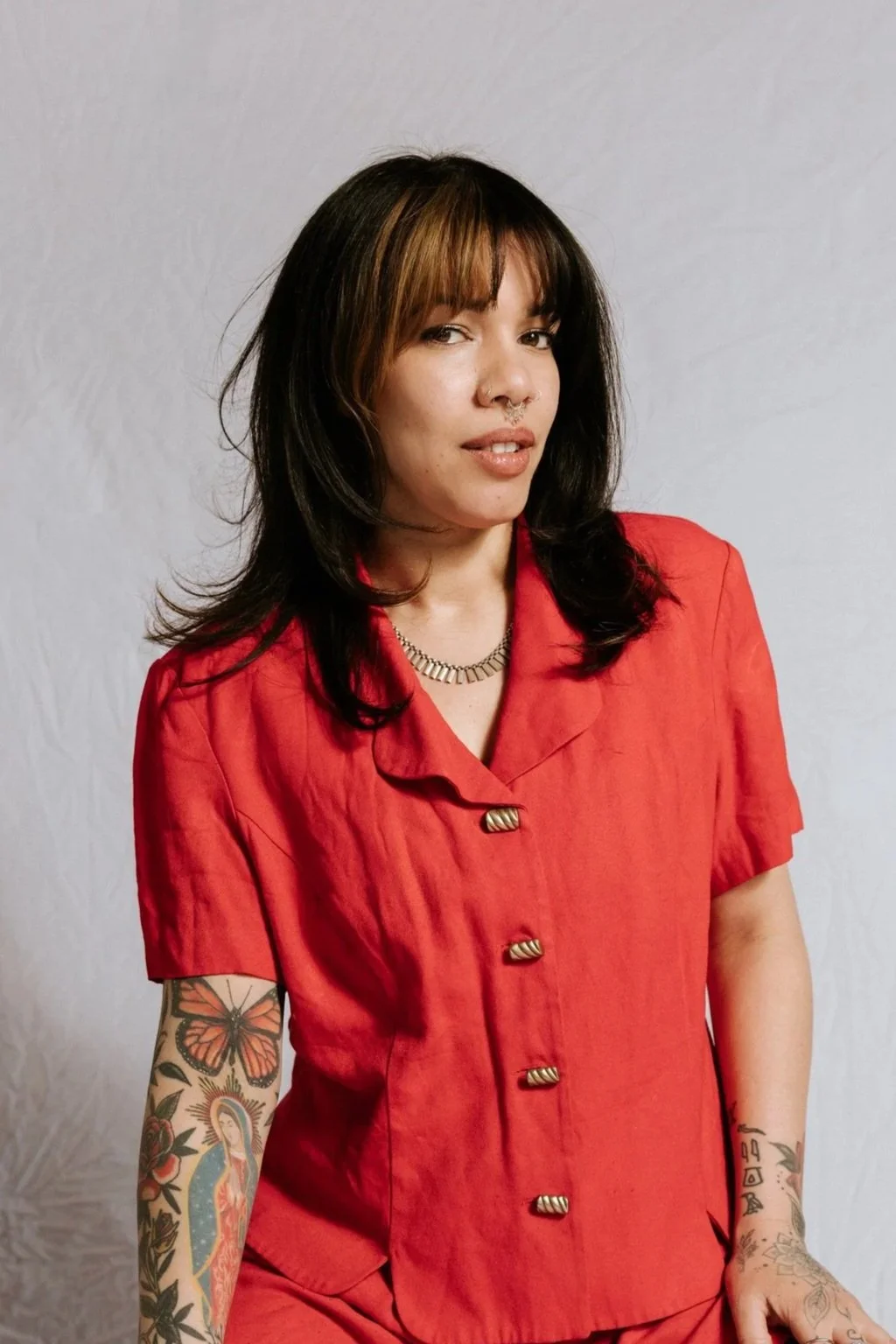 A young woman with dark hair and brown highlights wearing a red button-up shirt with short sleeves and a gold necklace, standing against a plain white background.