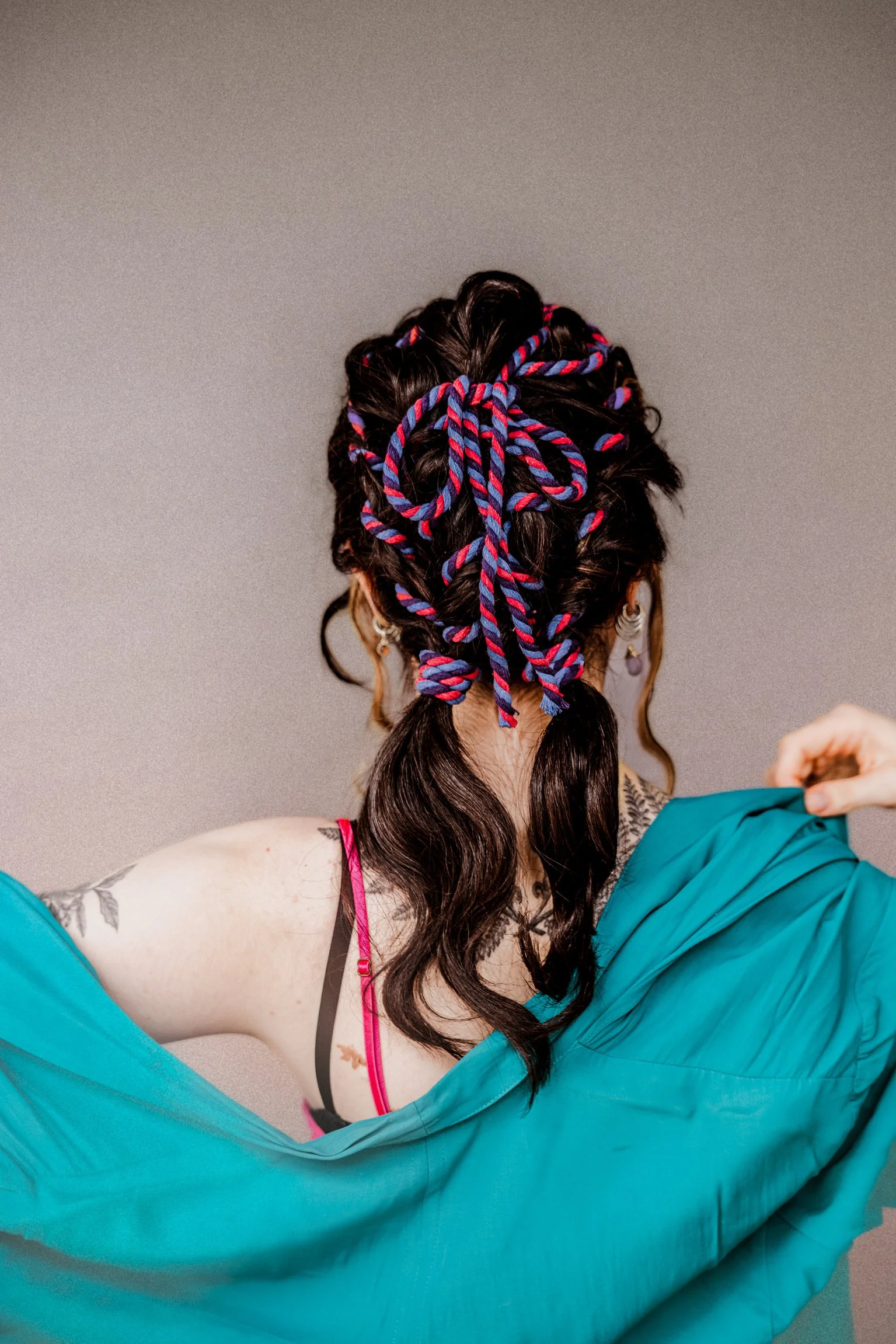 A woman with long dark hair decorated with multicolored braided hair extensions, wearing a pink bra strap, indoors against a plain wall, with a tattoo on her upper arm, and a turquoise jacket slipping off her shoulder.