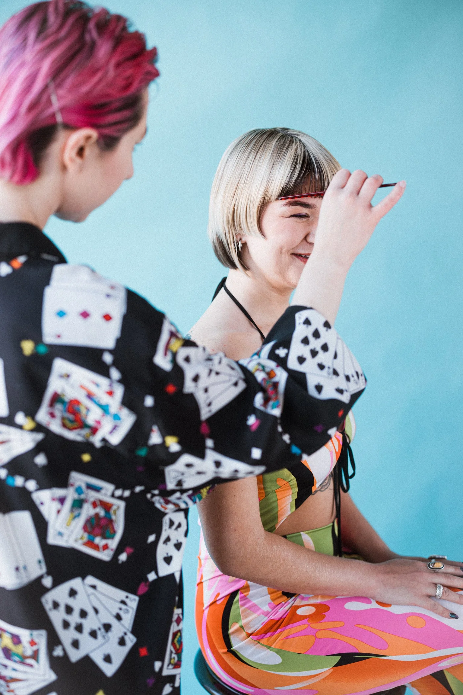 A woman with short blonde hair and a colorful dress sits on a turquoise chair, smiling, as another woman with pink and purple hair and a playing card-patterned jacket presents her with a tarot card or similar object. The background is a plain light blue color.