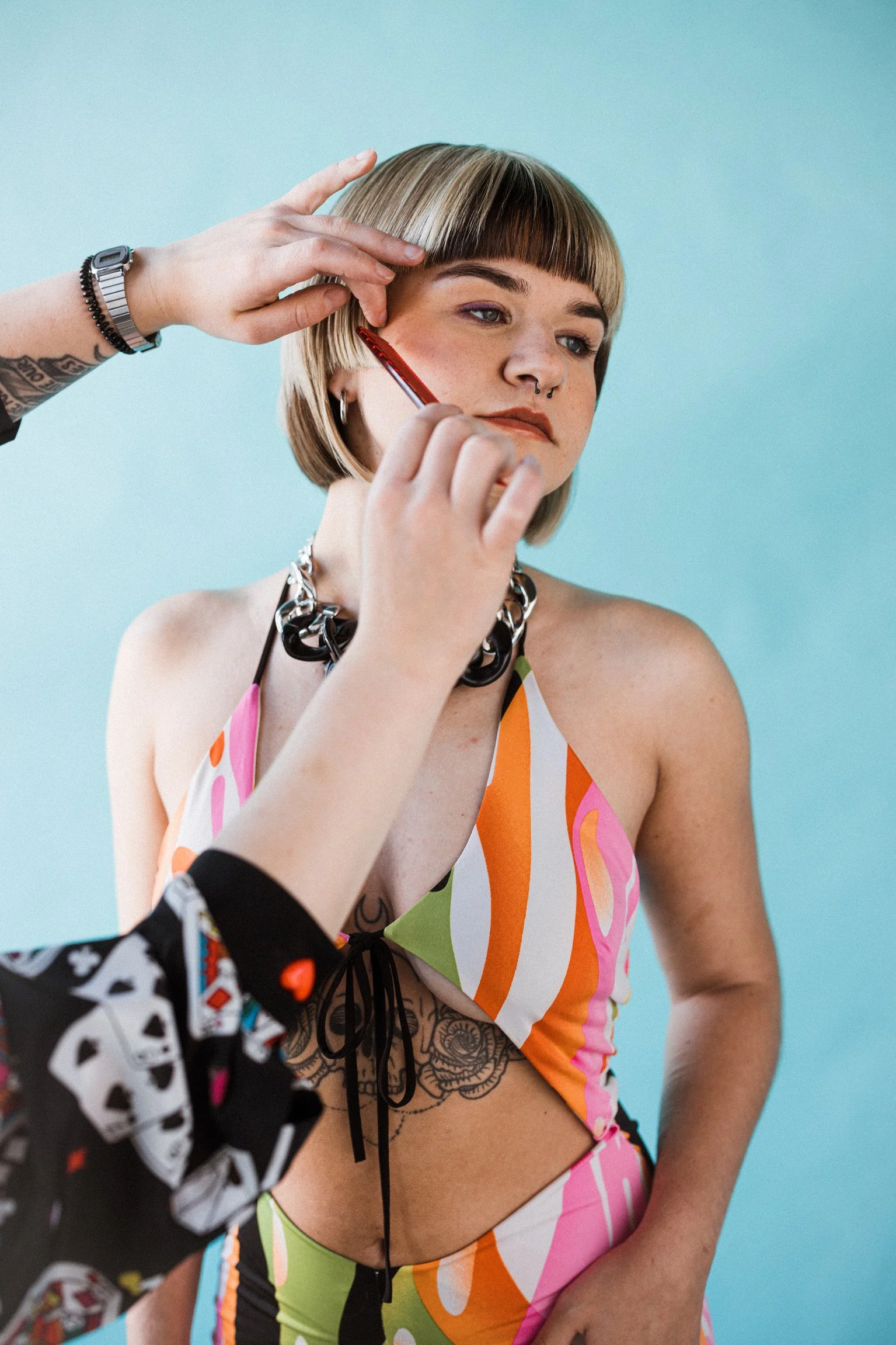 A woman with short blonde hair and a septum piercing is getting makeup applied to her face by another person. She is wearing a colorful, patterned outfit with orange, pink, green, and white. The background is turquoise.