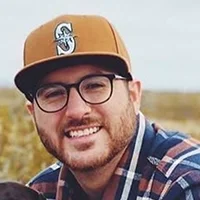 Professional headshot of a man wearing glasses and a tan baseball cap, smiling outdoors with a softly blurred natural background.