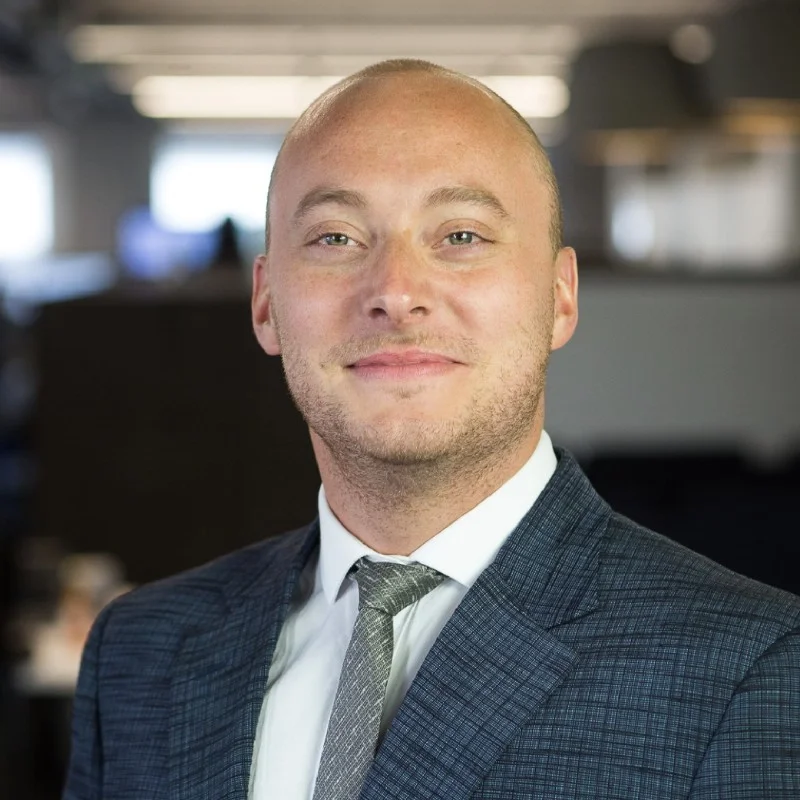 Professional headshot of a man in a blue suit and gray tie, photographed in a modern office with a softly blurred background and natural lighting.