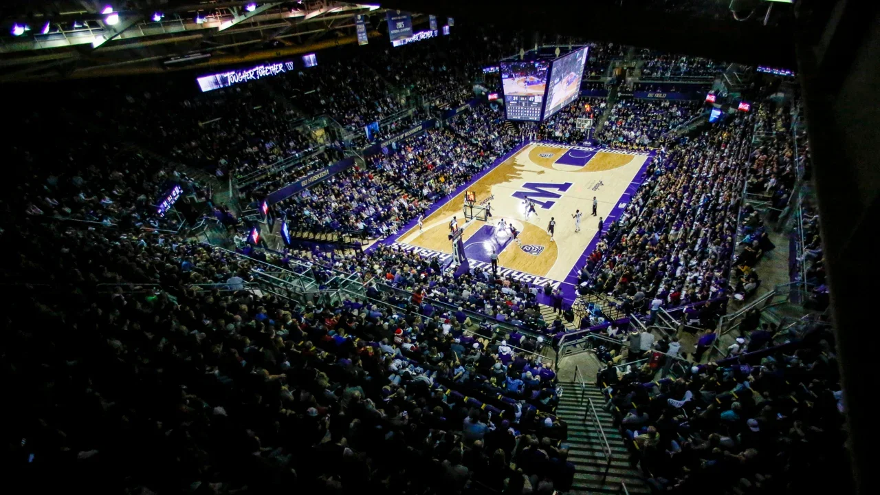 A packed indoor basketball stadium with fans seated around a basketball court with purple and gold markings, a large digital scoreboard hanging above, and personal cameras capturing the game.