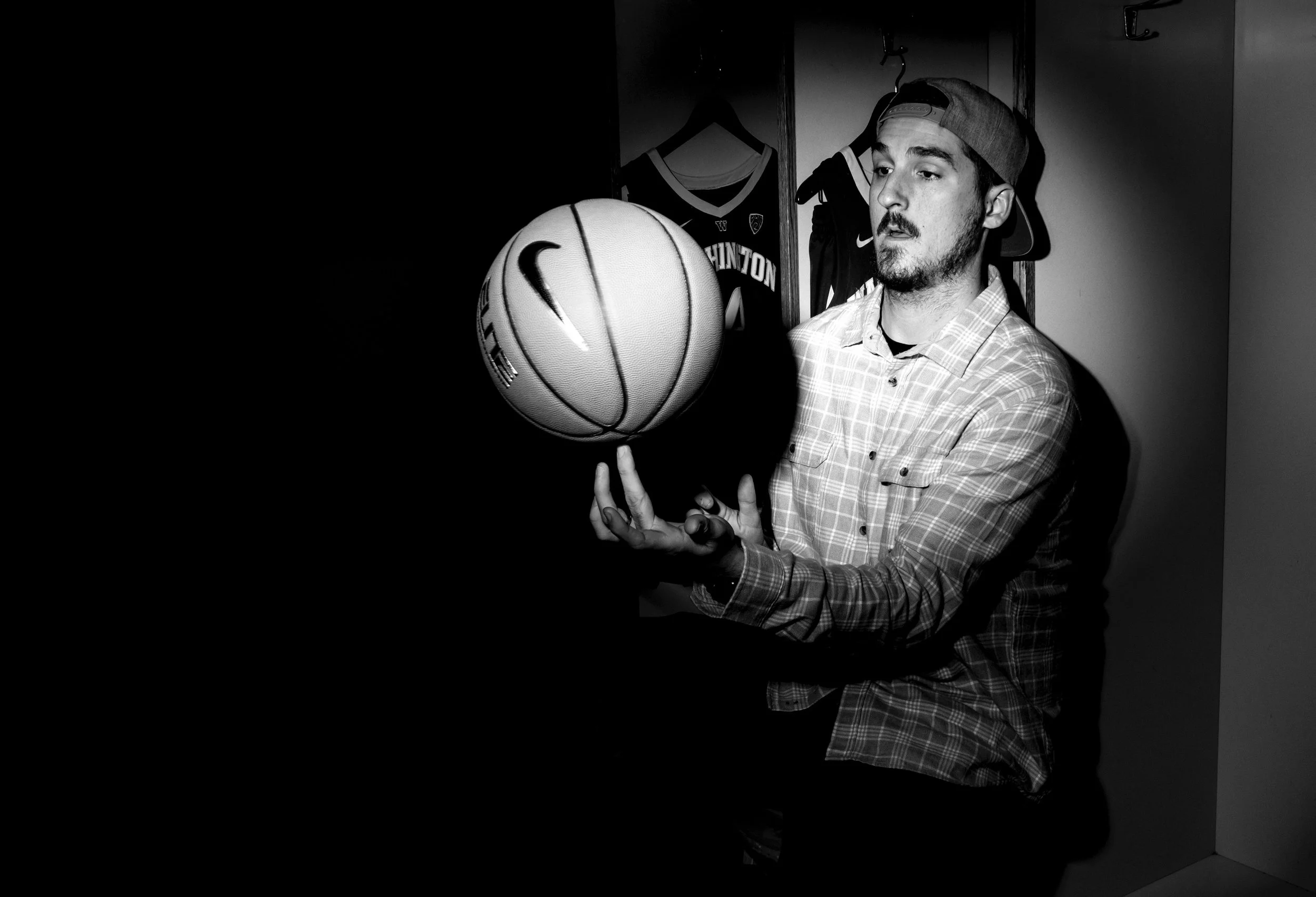Black and white photo of a man wearing a baseball cap backwards, holding a basketball with Nike logo, standing in a room with sports jerseys hanging in the background.