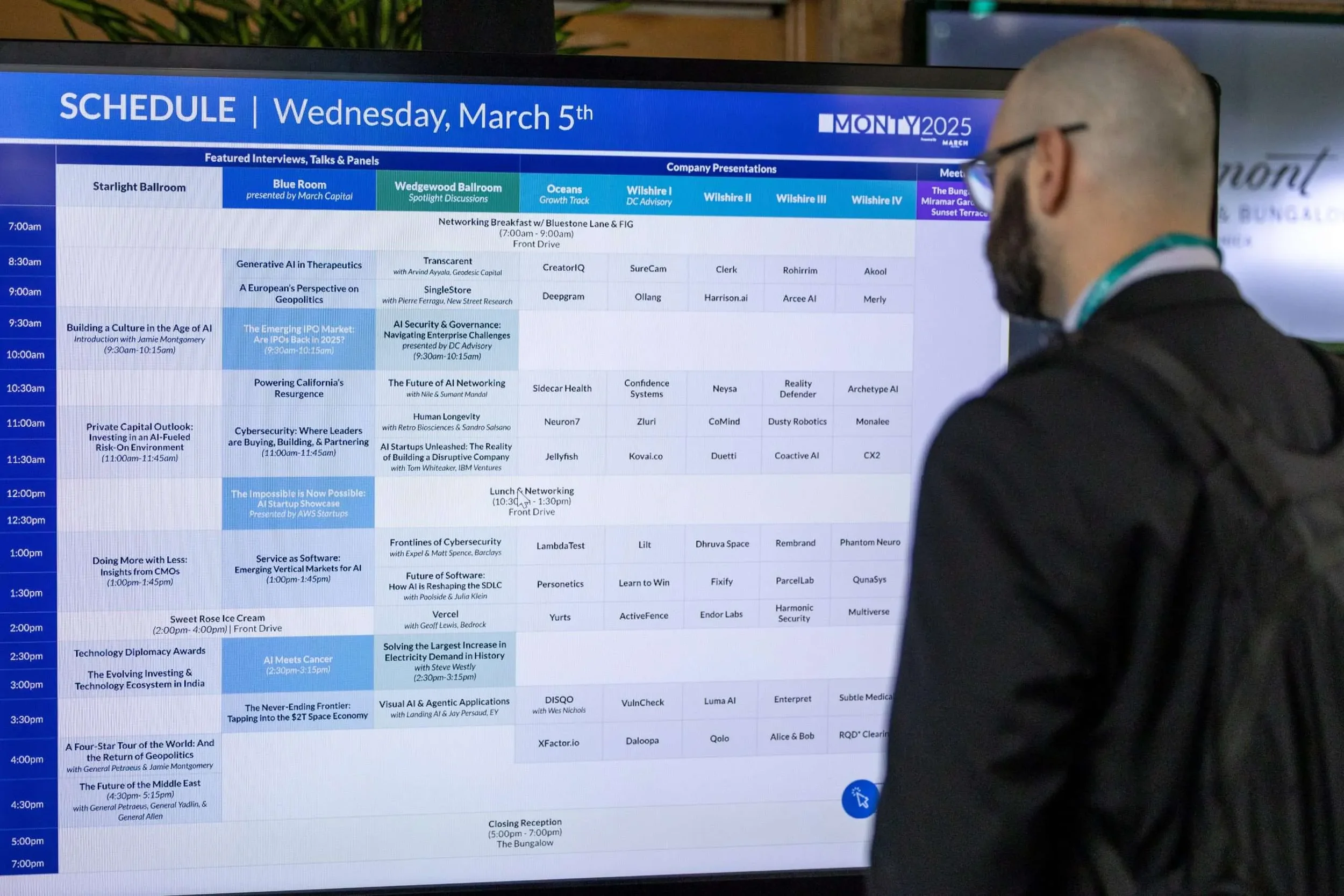 A man with a beard, glasses, and a bald head is looking at a large digital schedule board for a conference. The schedule displays events for Wednesday, March 5th, 2025, with sessions and times listed in a blue and white layout.