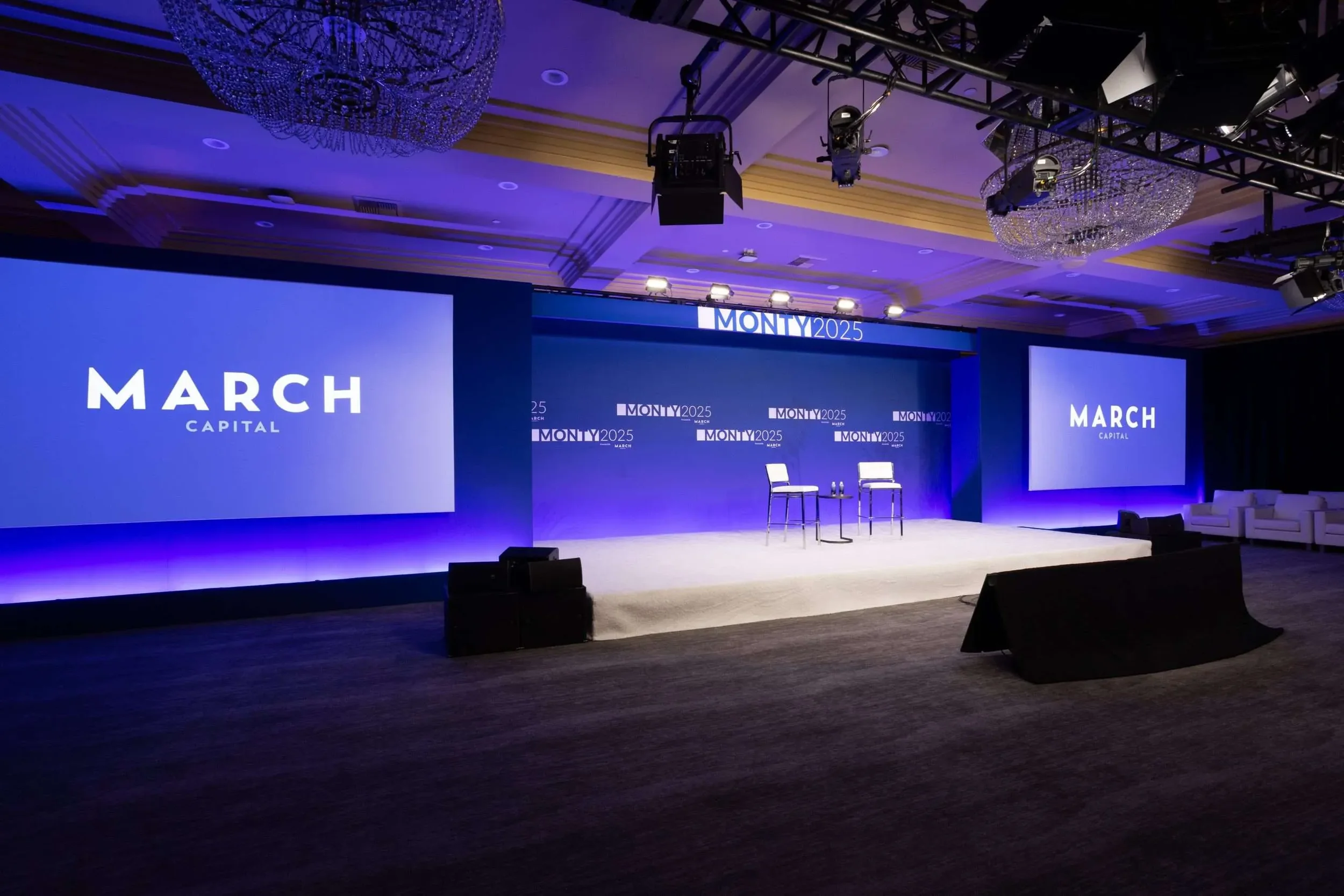 Empty stage setup with two chairs and a small table, large screens displaying 'MARCH CAPITAL', and a conference backdrop with 'MONTY2025' branding in a dimly lit, professional event hall.