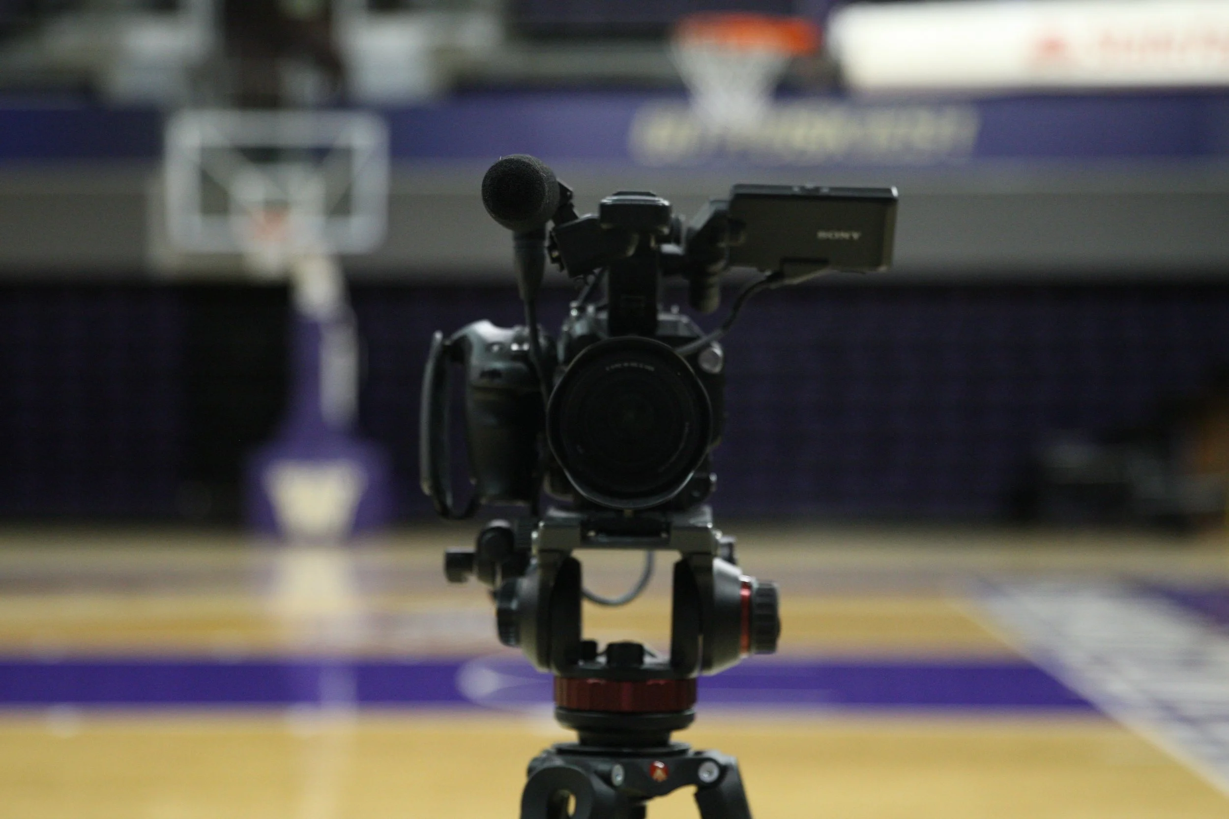 A professional video camera mounted on a tripod on a basketball court with purple and yellow lines, positioned to record a game, with blurred basketball hoop in the background.