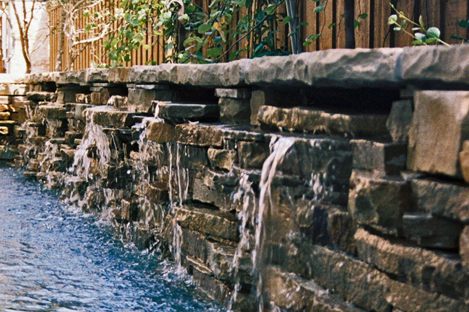A detail of a rustic, stone waterfall feature built on the back side of the pool to provide visual interest. Built by Ohlenburg Inc. Fine Swimming Pool Construction in Dallas, Texas.