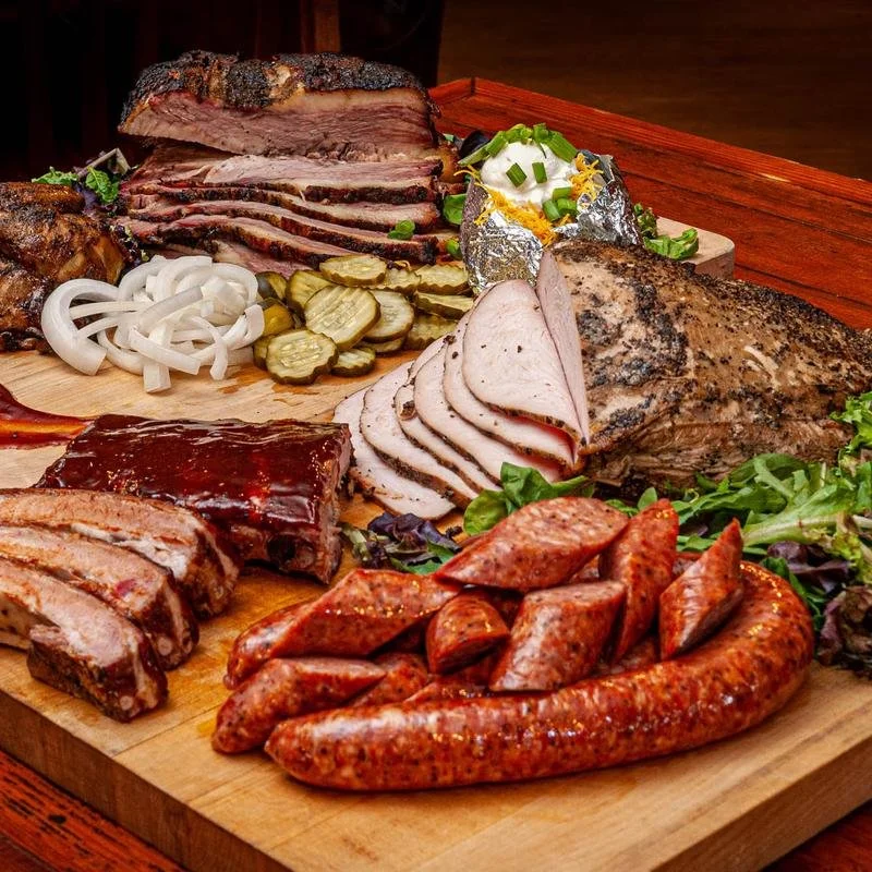 Assorted barbecue meats on a wooden platter including brisket, pulled pork, sausage, ribs, and sliced turkey, garnished with pickles, onions, and greens.