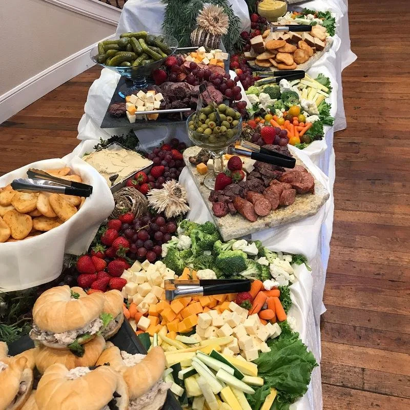 A buffet table with an assortment of cheeses, fruits, vegetables, pickup sandwiches, snacks, and pickles. The table is covered with a white tablecloth and decorated with greenery and flowers.