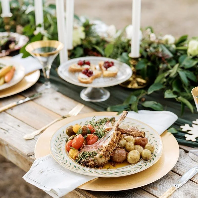 A holiday table setting with a plated meal of roasted lamb chop, cherry tomatoes, grapes, and herbs. In the background, there are festive decorations, candles, and a plate of cookies with icing and berries.
