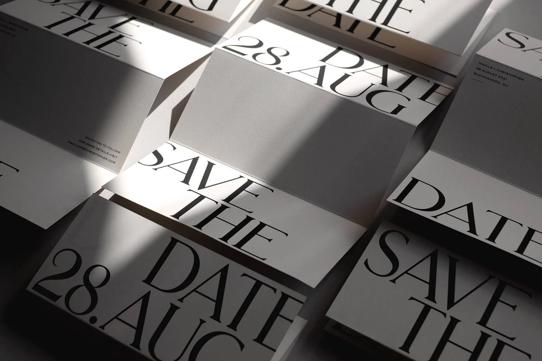 An angled view of unfolded Save the Dates resting on a table with light pouring in from a window on the right. The words SAVE THE DATE and 28.AUG are printed in large black ink across the top and bottom of each page with blank space in the middle.