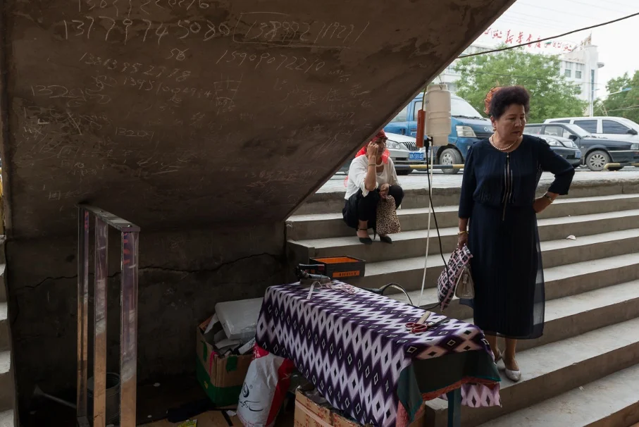 A seamstress uses the available space under a stairwell at a local bazaar. The client can wait to have the alterations or write down their phone number among those of other clients so she can call when it is complete. 