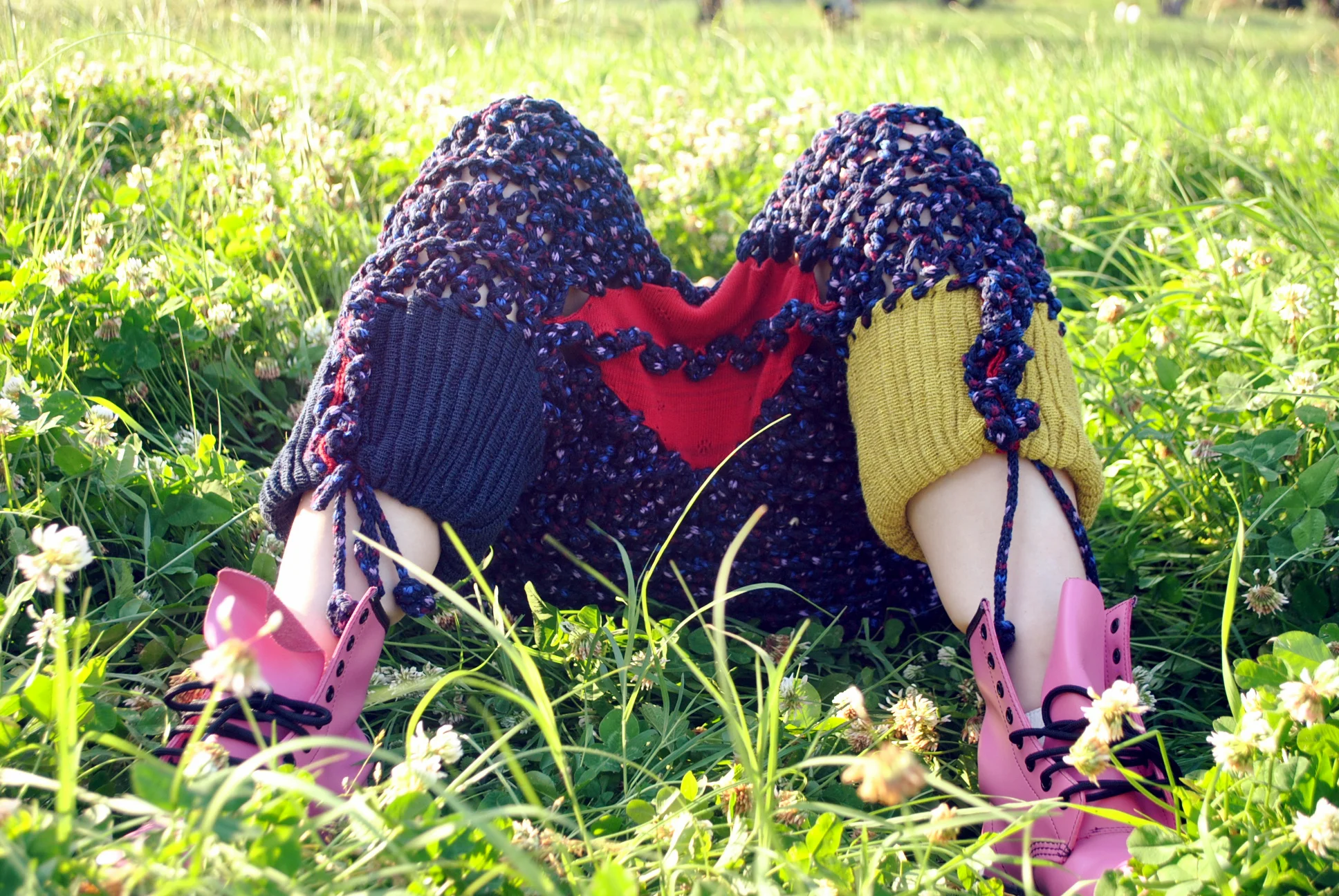 Child wearing pink rain boots sitting in a grassy field with white flowers, surrounded by green foliage and sunlight.