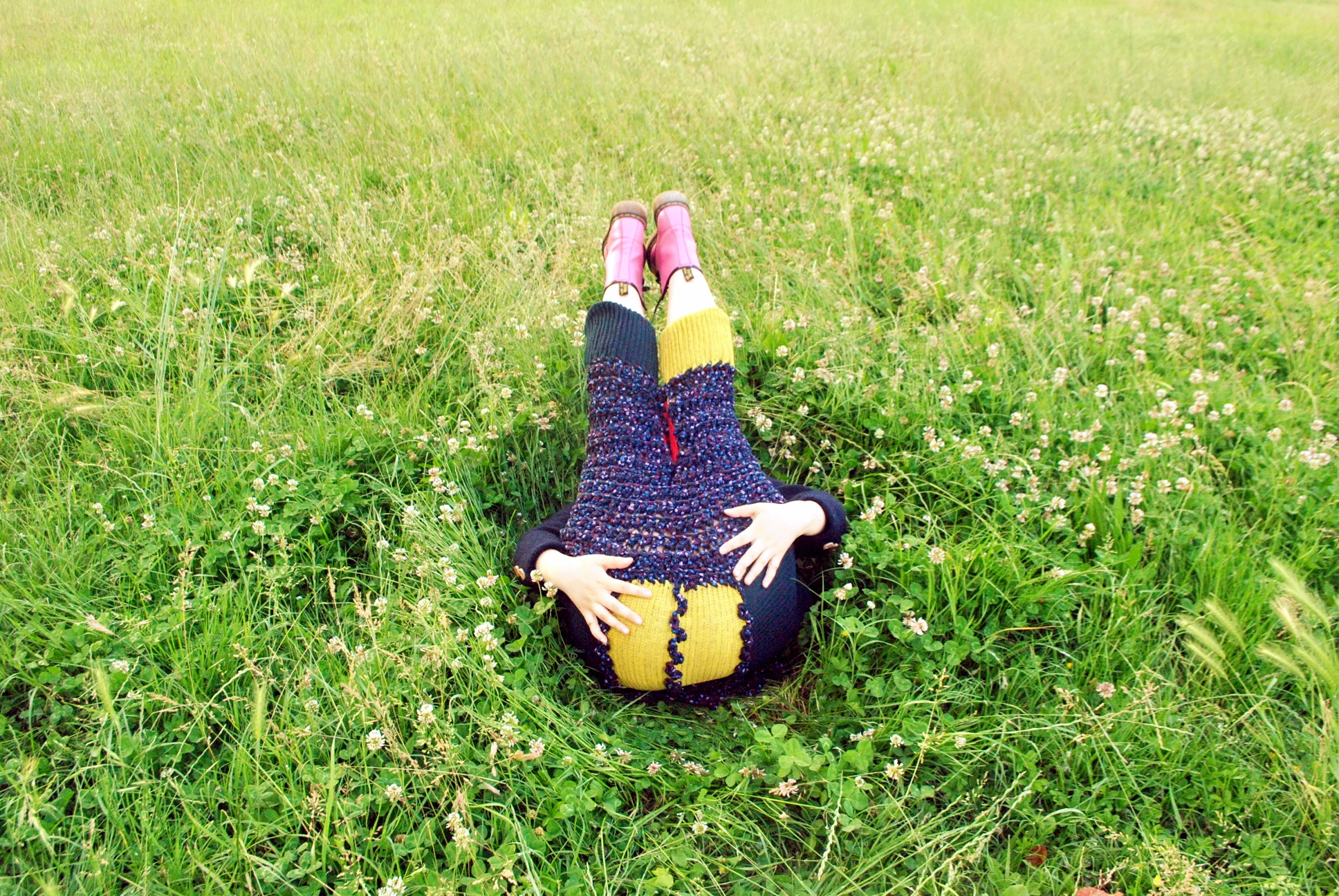 A person lying on the grass in a field of small white flowers, wearing a colorful sweater, yellow and black pants, and pink boots.