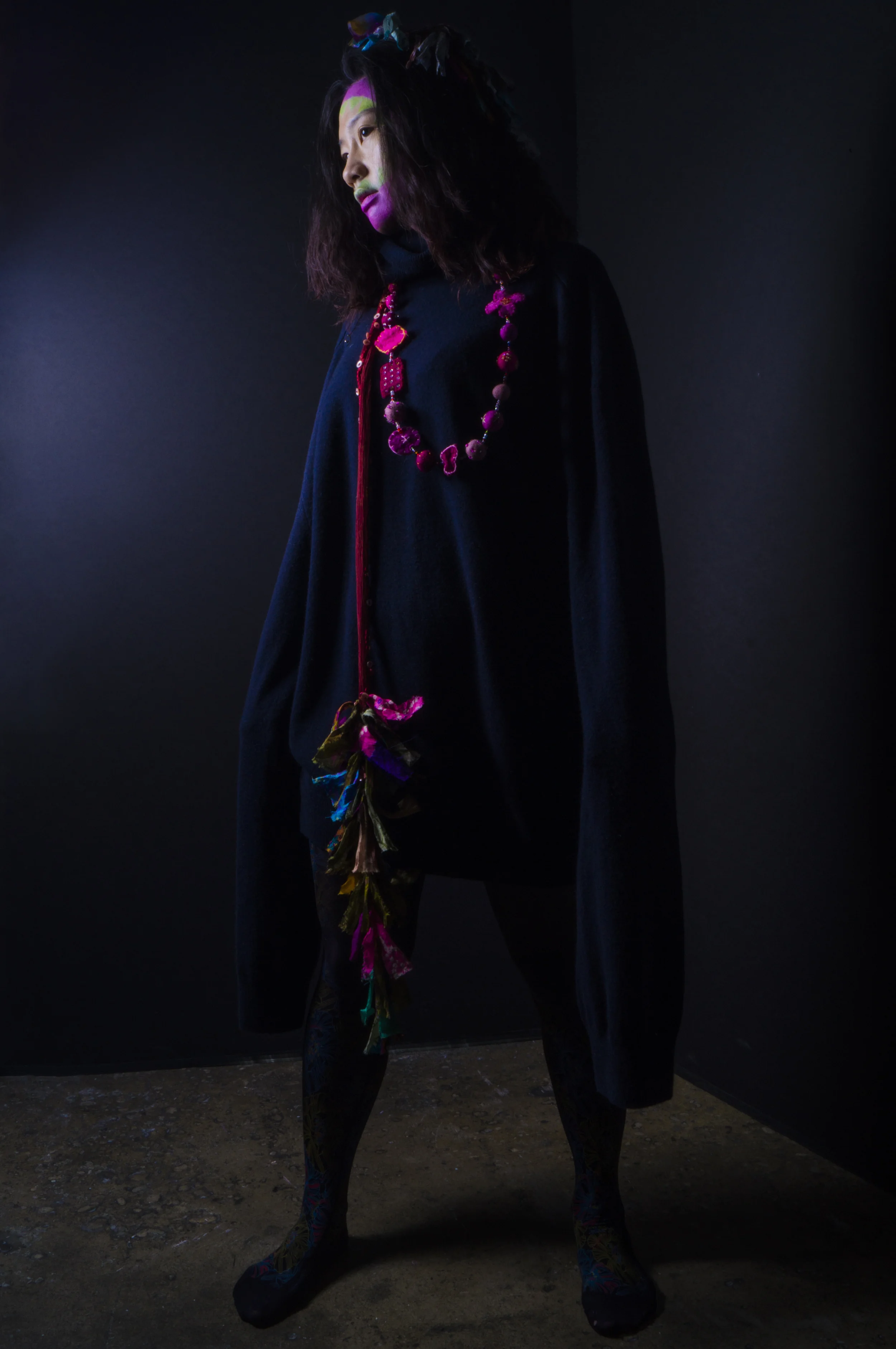 A woman with dark hair in loose waves, wearing colorful makeup, a black overcoat with vibrant accessories, standing on a dark, textured floor in front of a black background.